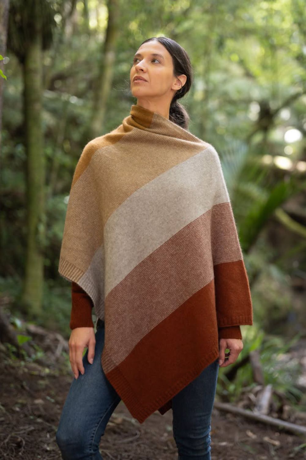 A woman wearing the Ombre Possum Merino Poncho in Harvest by McDonald, standing in a lush green forest.