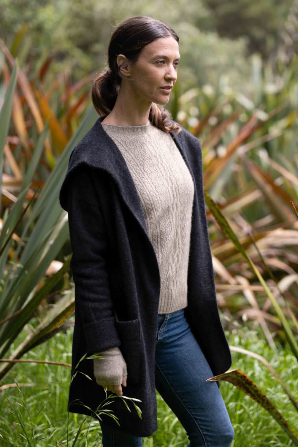 A woman wearing the McDonald Oversize Hood Possum Merino Jacket in Charcoal over a cream cable-knit sweater and blue jeans, standing outdoors in a grassy, leafy environment.