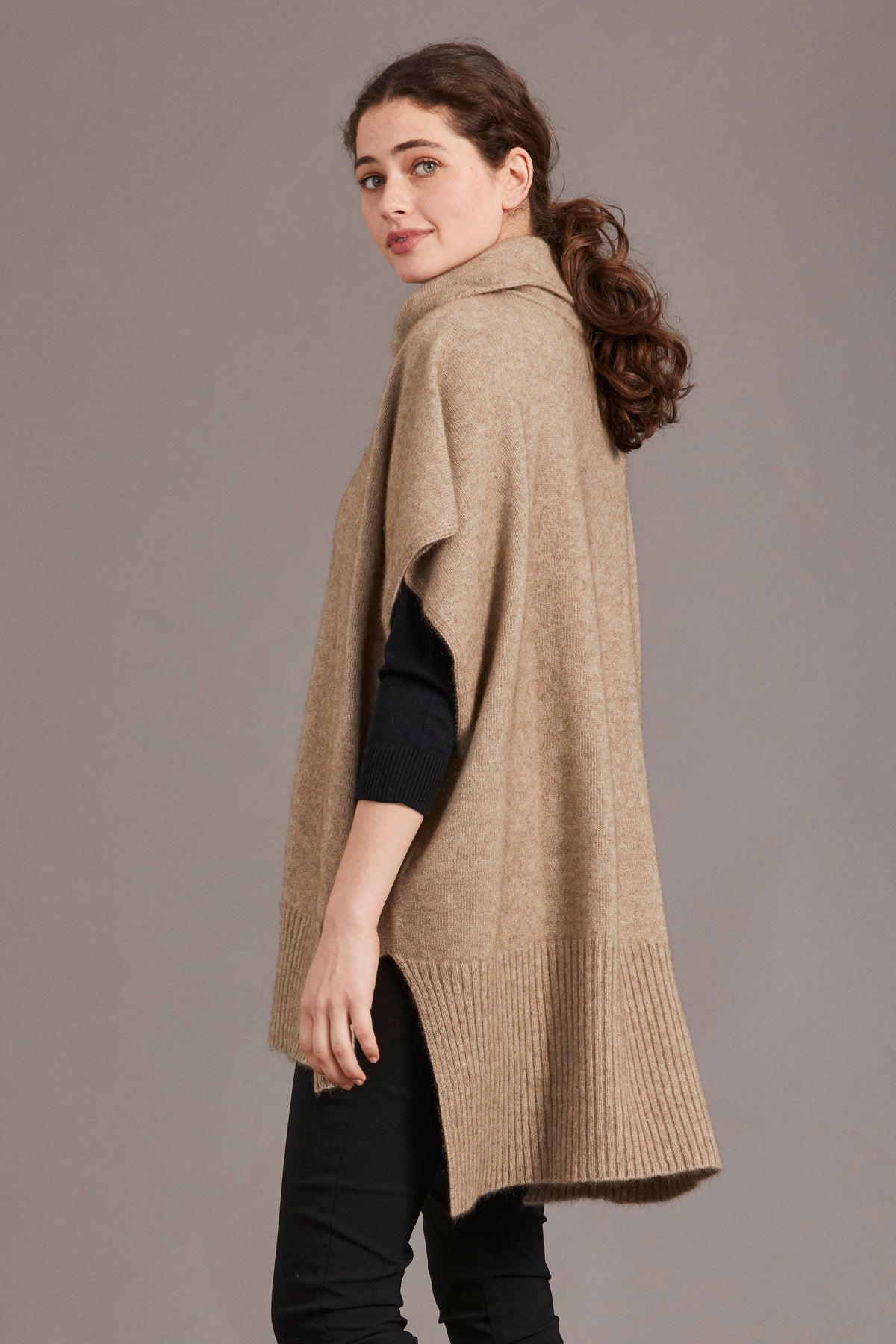 Woman wearing the Step Hem Possum Merino Poncho in Mocha by McDonald against a plain background - side view