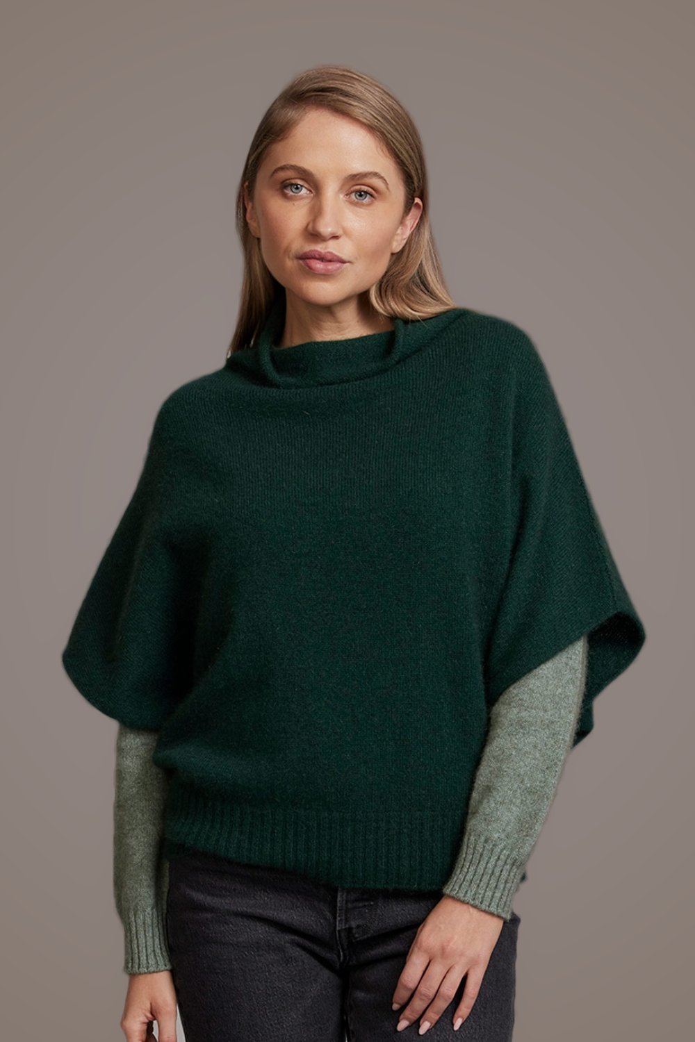 Woman wearing a Possum Merino Shrug Style Sweater in Forest by McDonald layered over a mint green sweater, with a plain background