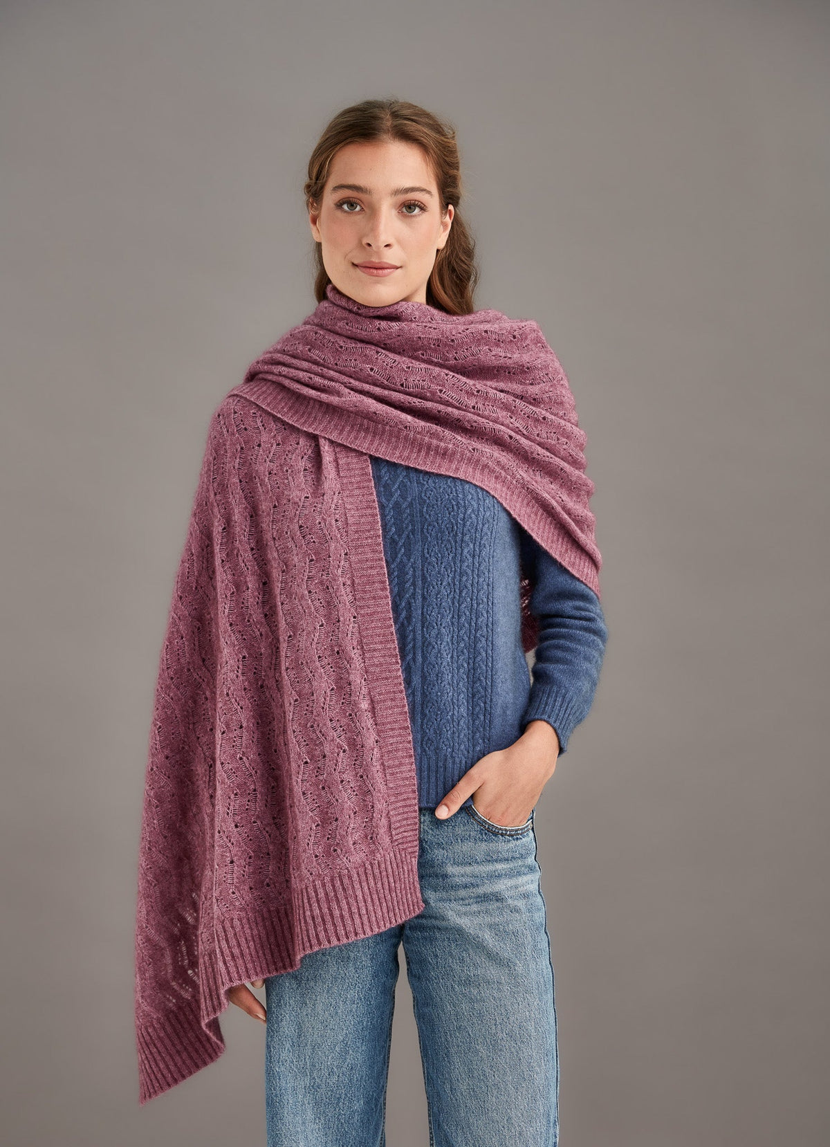 Woman wearing a pink knitted shawl over a blue sweater and jeans on a gray background