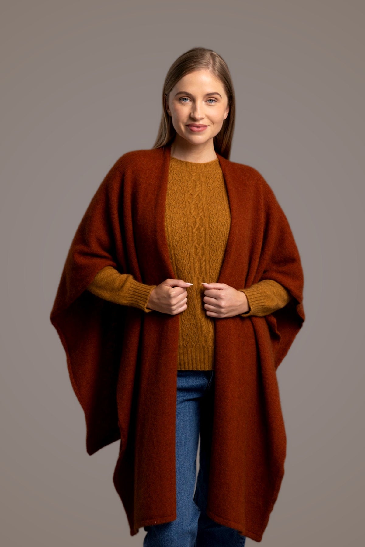 Woman wearing the Split Front Possum Merino Wrap in Spice by McDonald over a gray sweater and blue jeans on a brown background