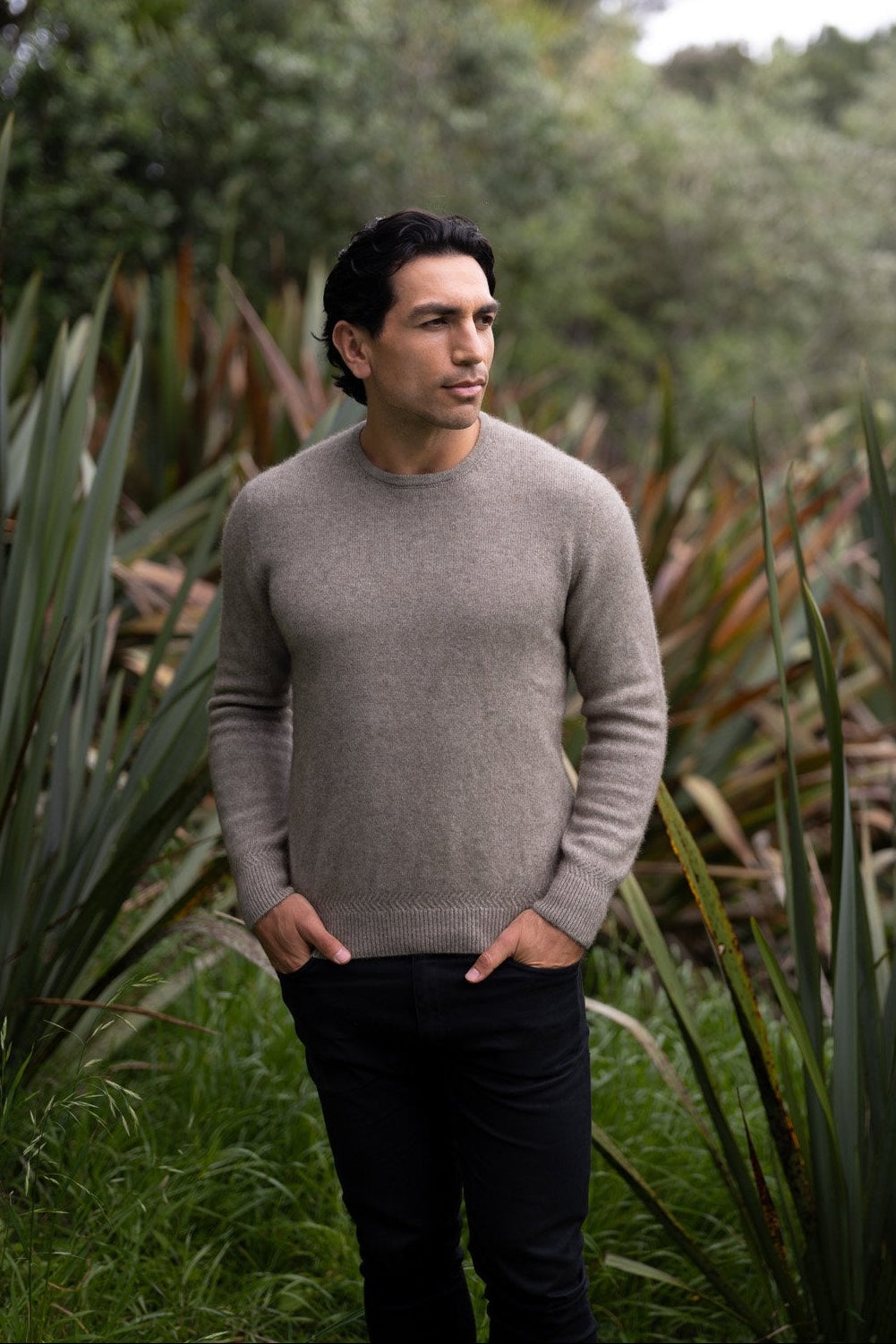 Man wearing the Rack Stitch Crew Neck Possum Merino Jersey in Mocha by McDonald standing among green plants