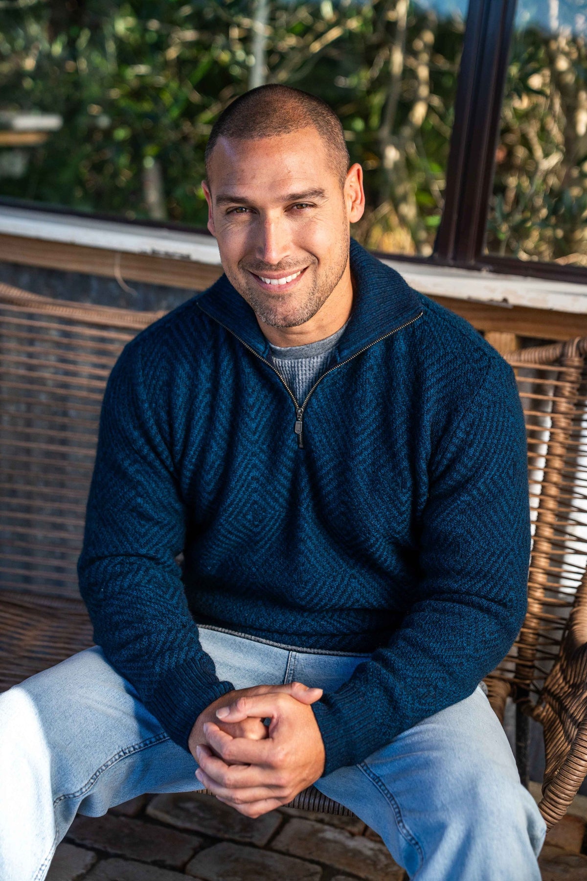 A smiling man with a buzz cut sits on a wicker bench wearing the Diamond Plated Zip Possum Merino Jumper in Navy/Teal by Brushtail over a gray t-shirt and light blue jeans.