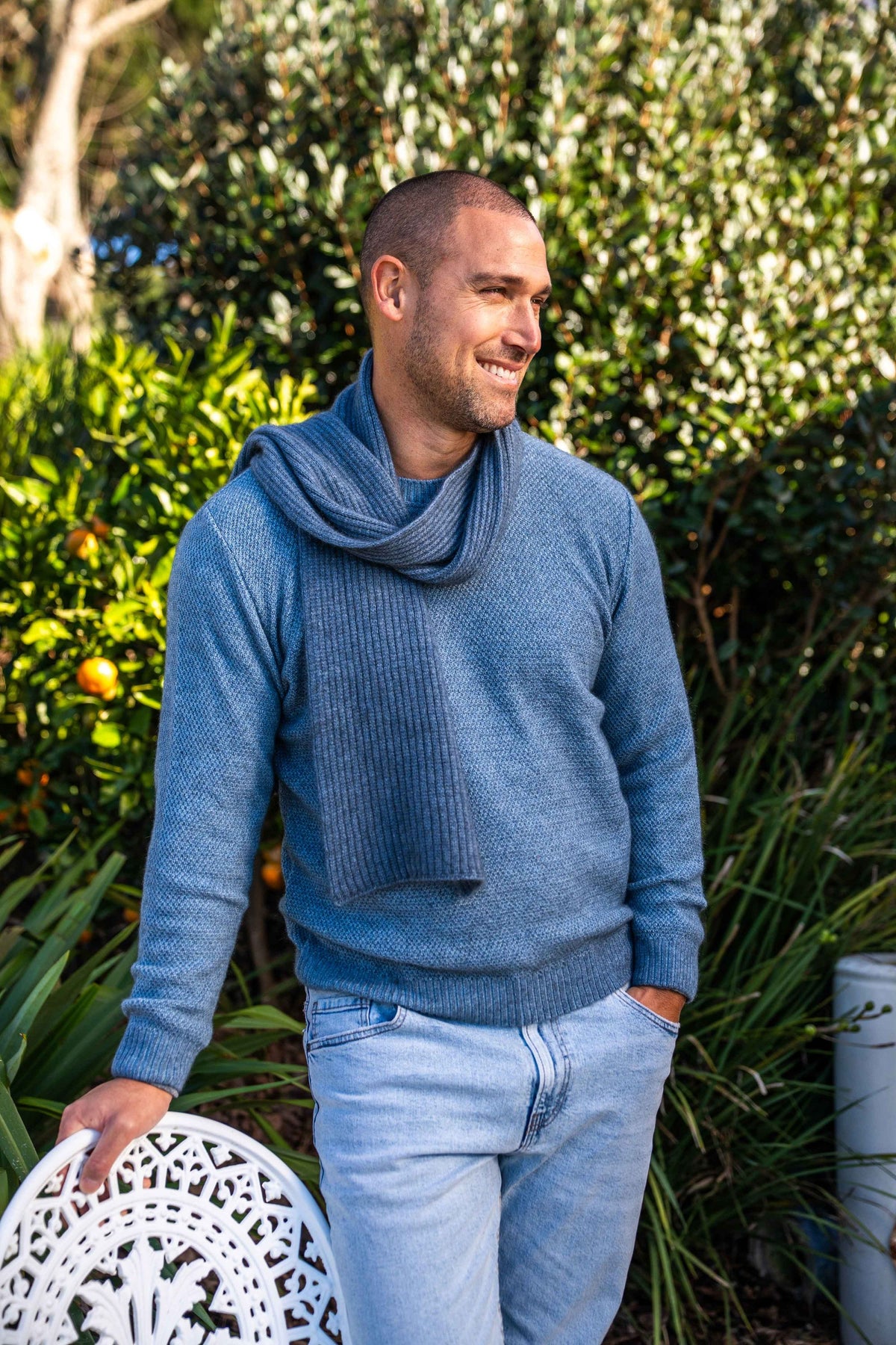 A man wearing the Moss Stitch Plated Possum Merino Jumper in Riviera/Sky by Brushtail by Koru and matching blue knit scarf, standing outdoors in front of green foliage and holding a white decorative chair.