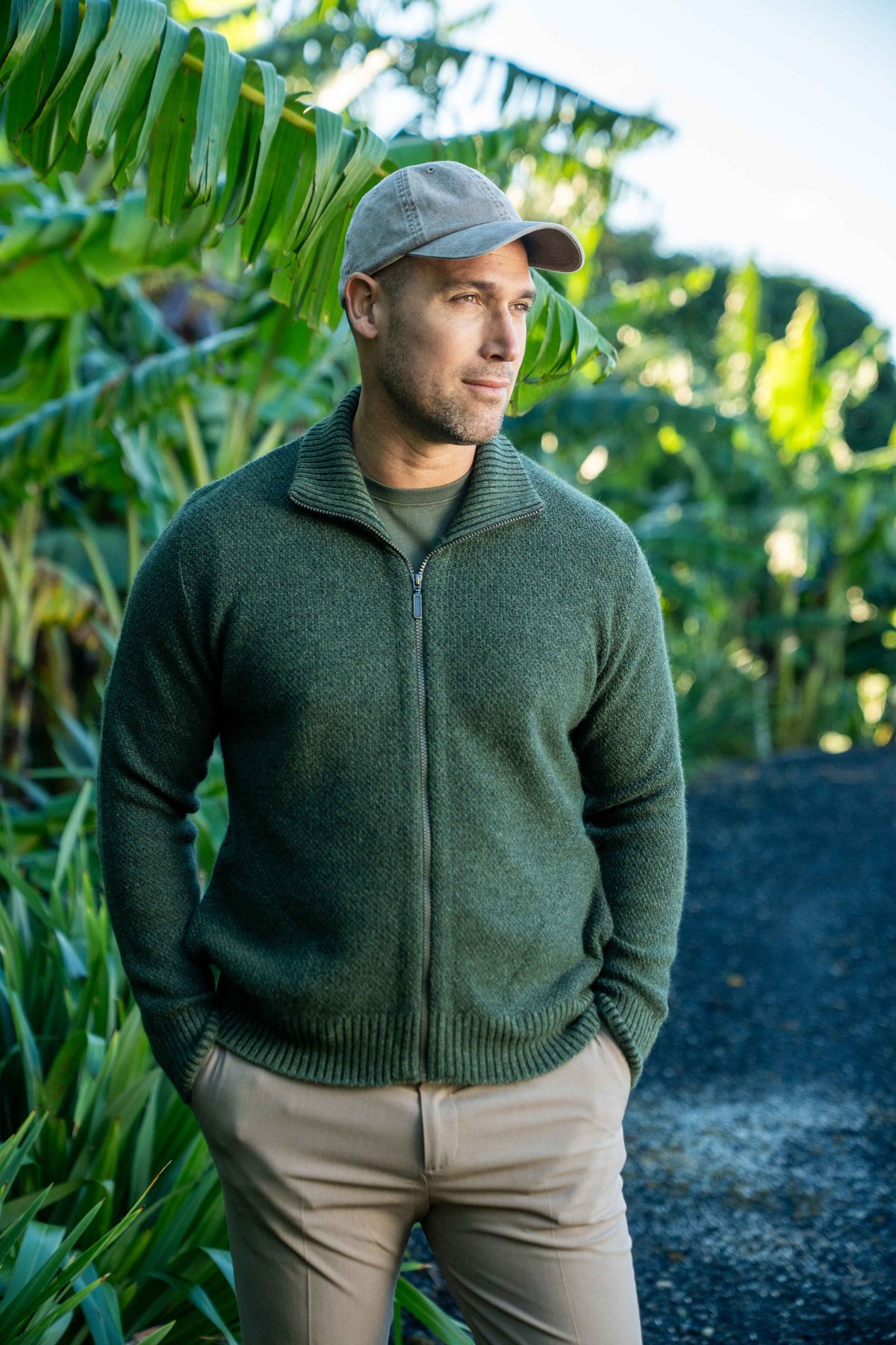 A man wearing the Moss Stitch Plated Possum Merino Jacket in Moss/Dark Green by Brushtail by Koru, khaki pants, and a gray cap, standing outdoors in front of lush green tropical foliage.