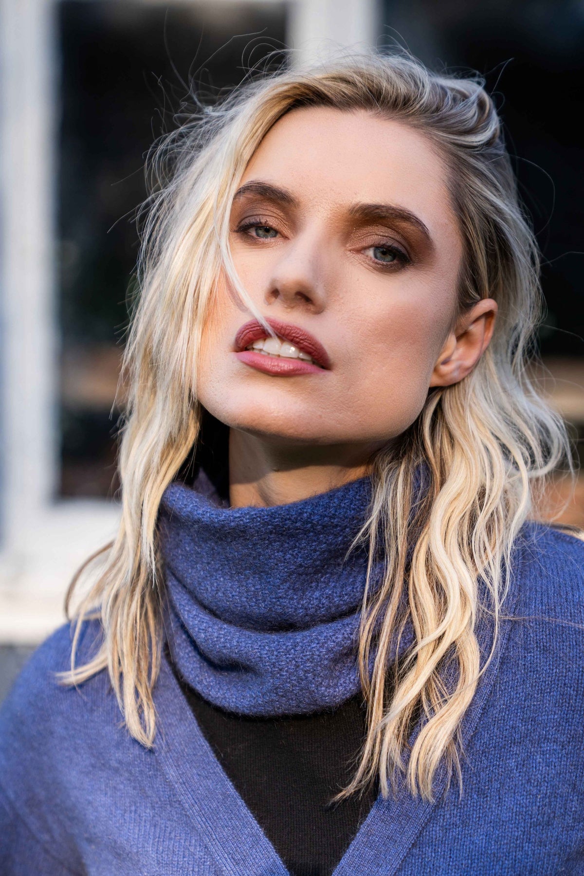 A close-up portrait of a blonde woman wearing the Moss Stitch Possum Merino Neck Warmer in Periwinkle by Koru over a black top, with a matching blue cardigan draped off her shoulders.