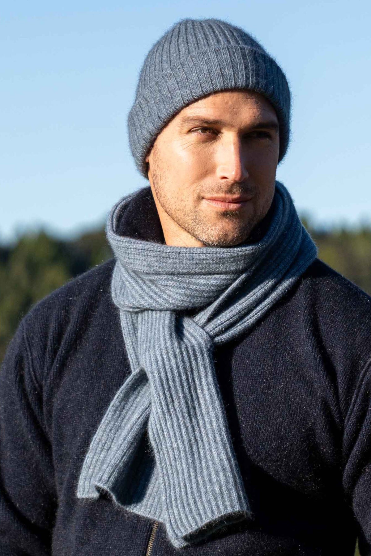 A man wearing a ribbed knit beanie and matching Ribbed Keyhole Possum Merino Scarf in Riviera by Koru, photographed outdoors against a soft blue sky and blurred trees.