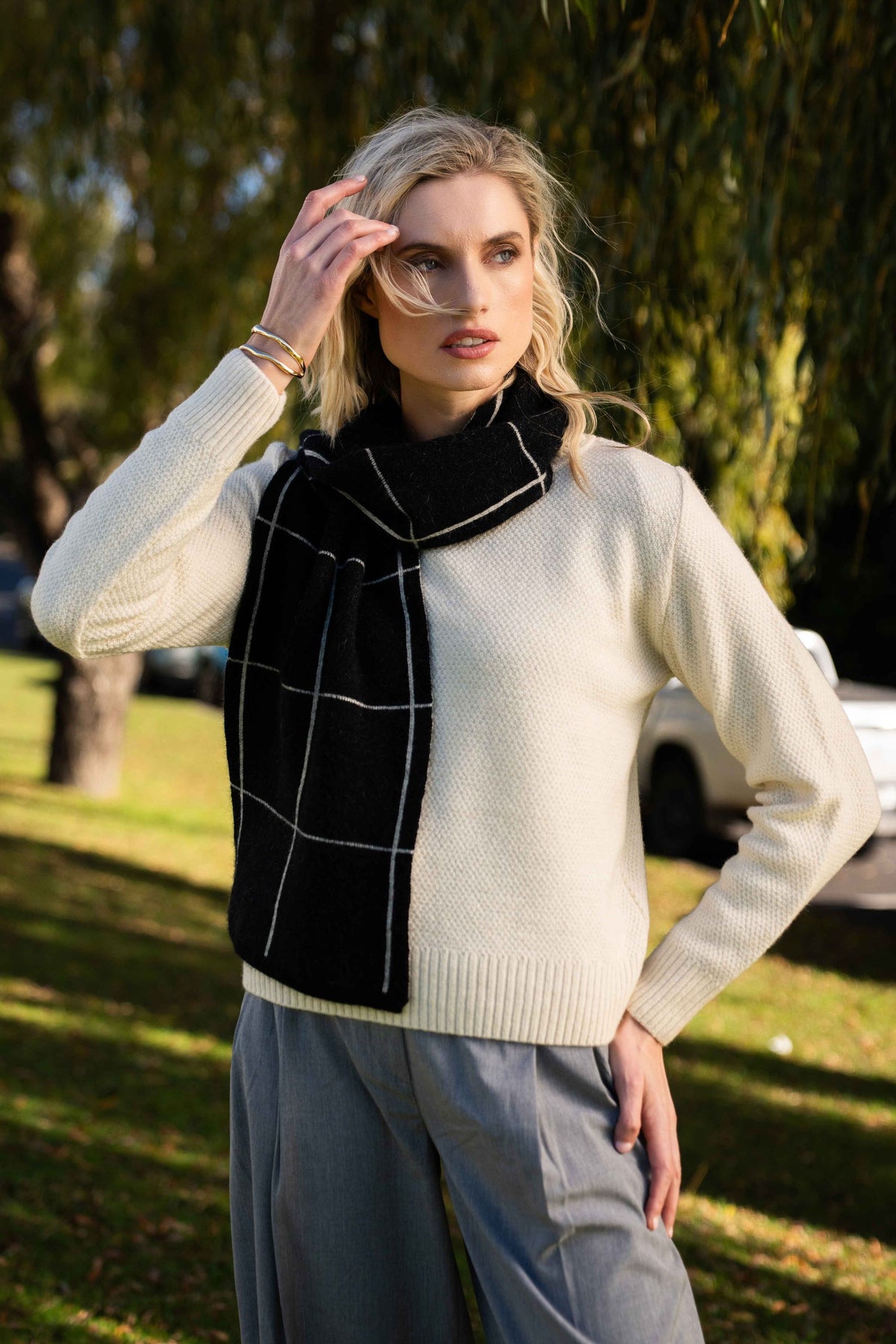 A woman outdoors wearing a cream knit sweater, gray wide-leg pants, and the Check Possum Merino Scarf in Black/White by Koru