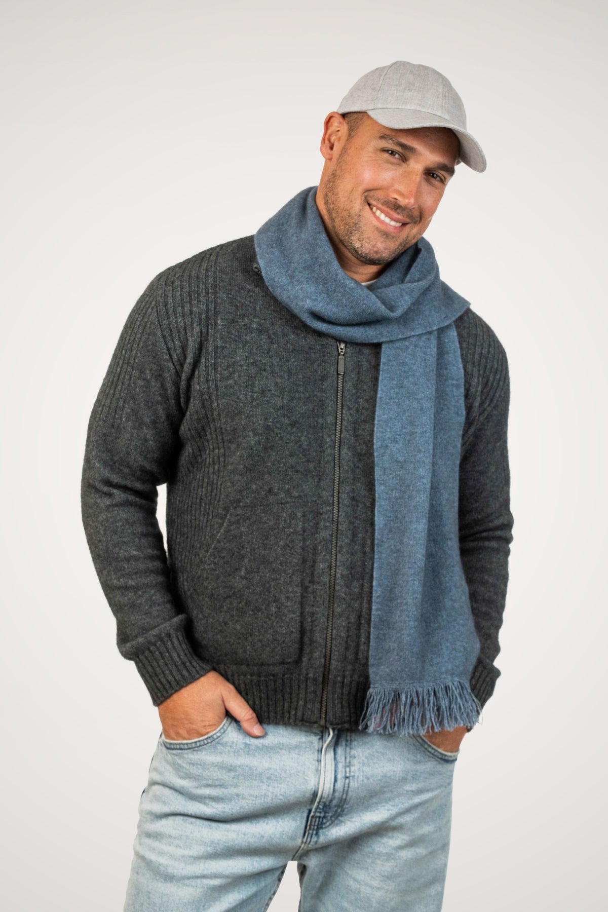 A man wearing a dark gray ribbed zip-up sweater, the Fringed Possum Merino Scarf in Riviera by Koru and a light gray baseball cap, standing against a plain light gray background.