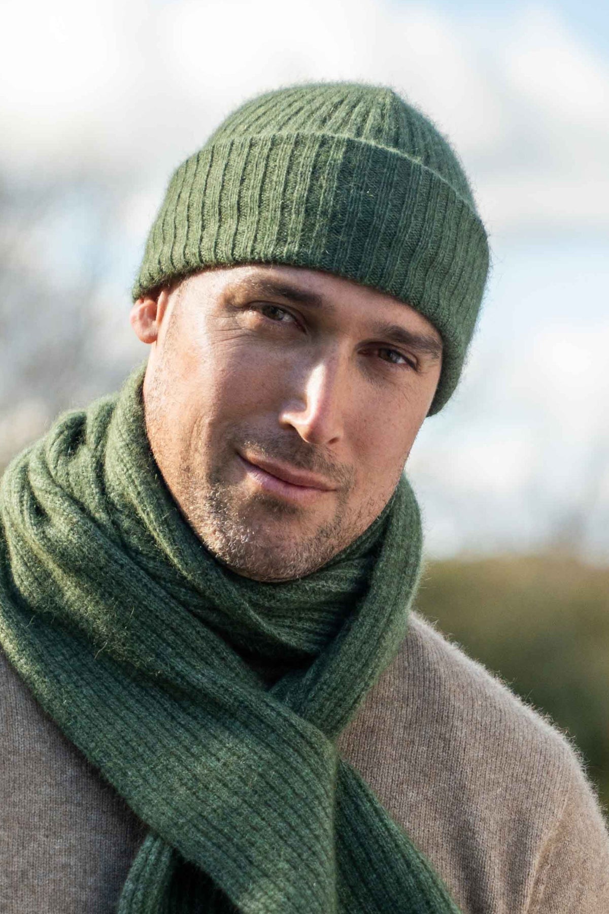 A man wearing the Lightweight Ribbed Possum Merino Beanie in Moss by Koru and matching knit scarf in olive green, with a beige coat, photographed outdoors against a softly blurred sky and trees.