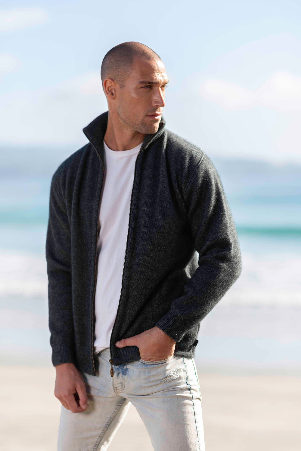 Man wearing the Zip Through Possum Merino Jacket in Grey by Koru over a white shirt with light blue jeans on a beach.