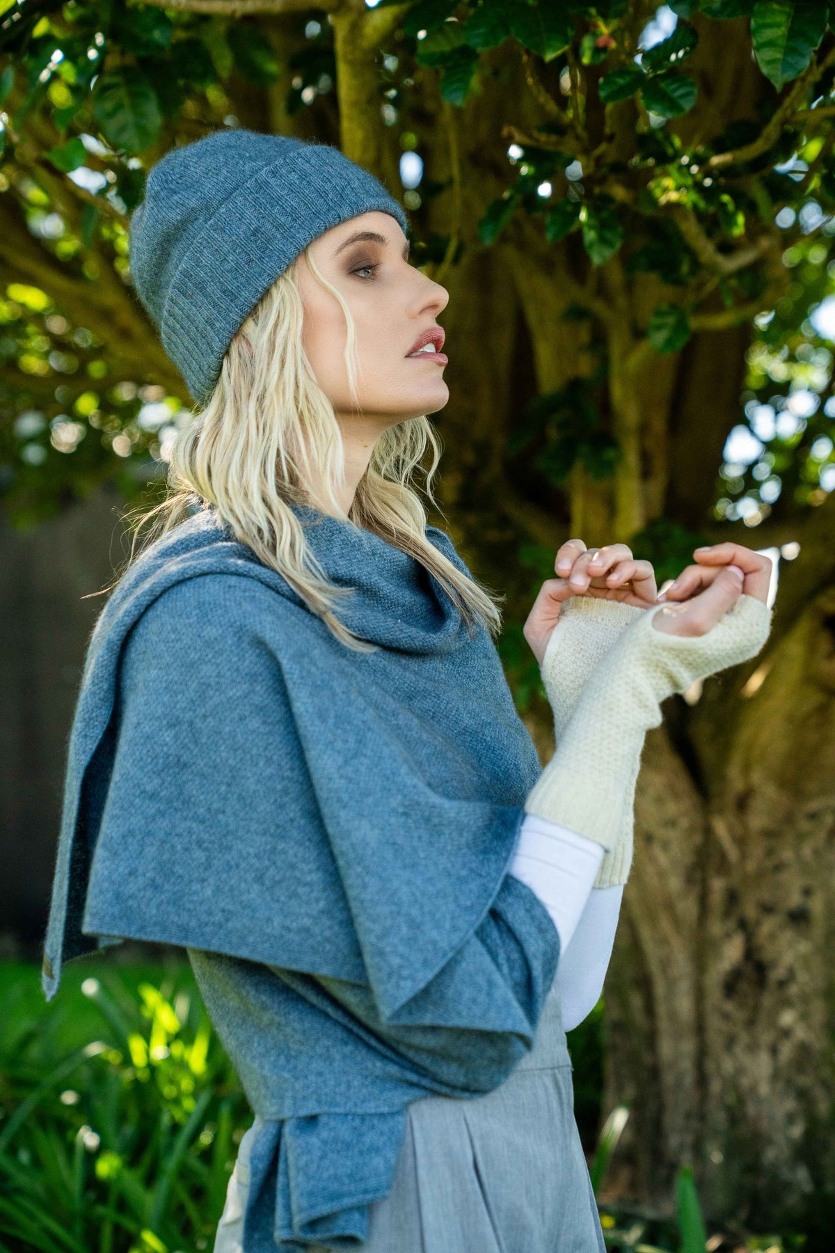 A woman outdoors wearing a blue knit beanie, matching the Moss Stitch Possum Merino Wrap in Riviera by Koru, and cream knit fingerless gloves, standing in front of a large tree.