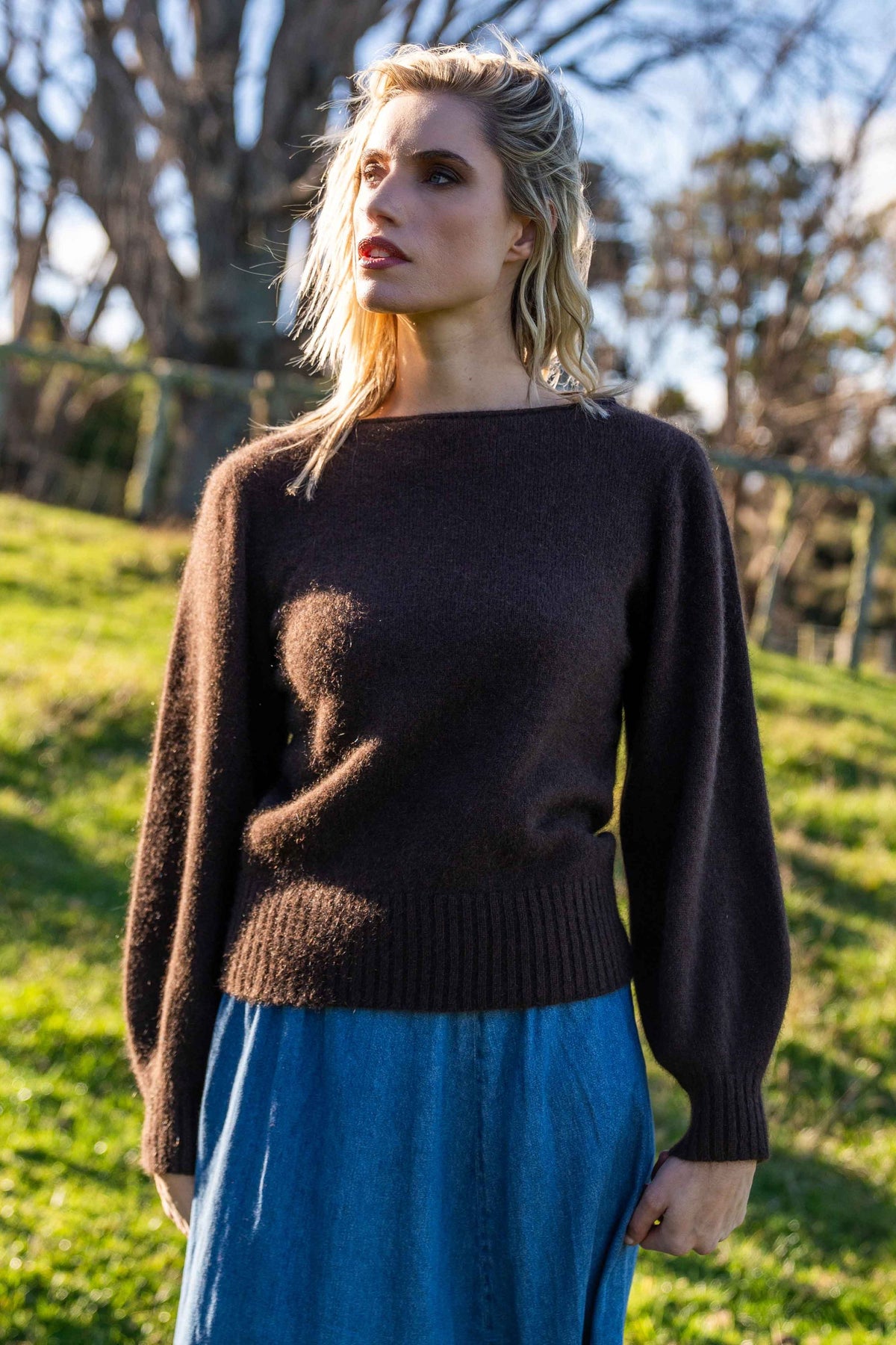 A woman wearing the Boat Neck Possum Merino Jumper in Chocolate by Koru and a blue skirt standing outdoors on a grassy hill with trees in the background.