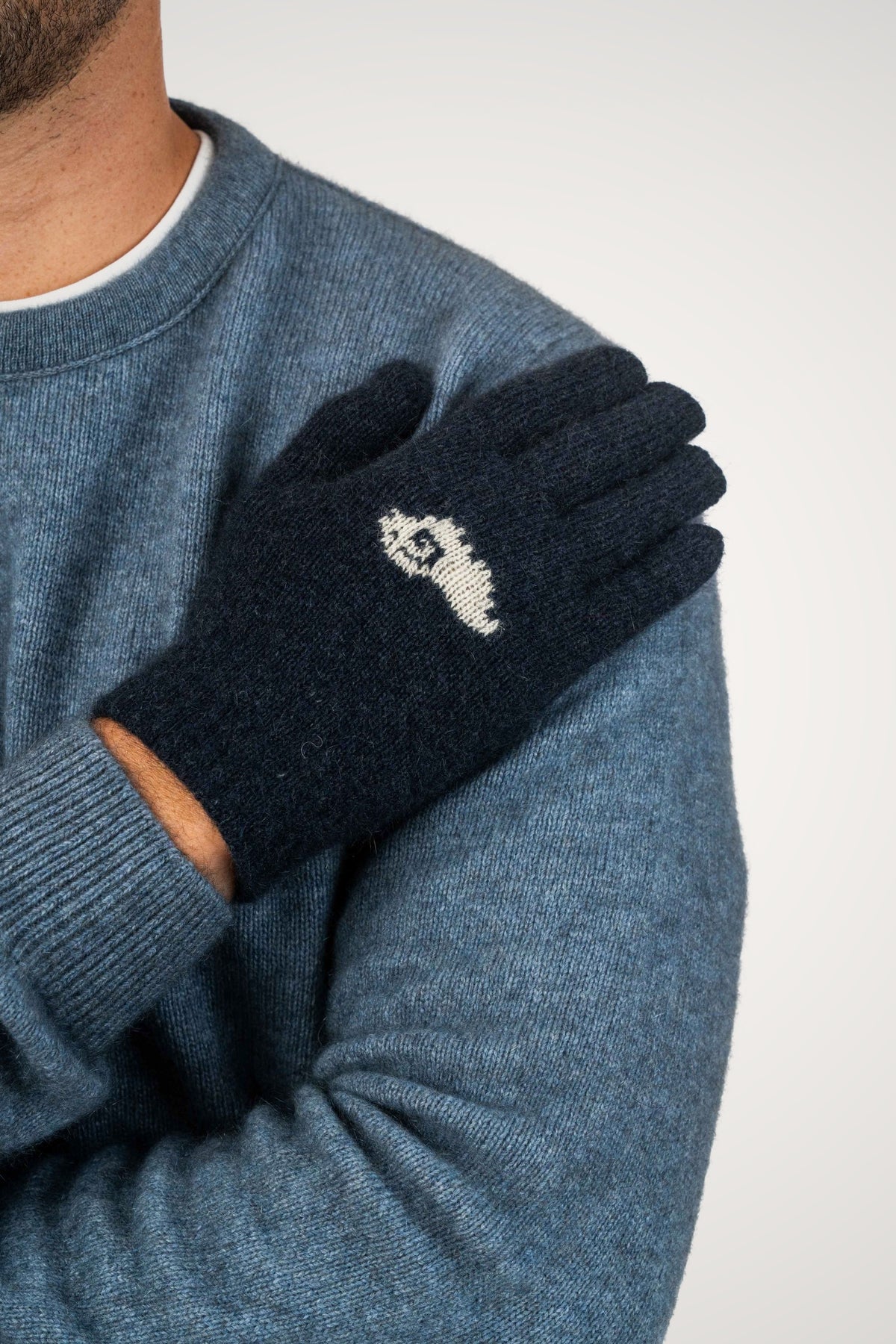 A close-up of a person wearing the Sheep Possum Merino Gloves in Indigo by Koru, paired with a blue-gray knit sweater against a light gray background.