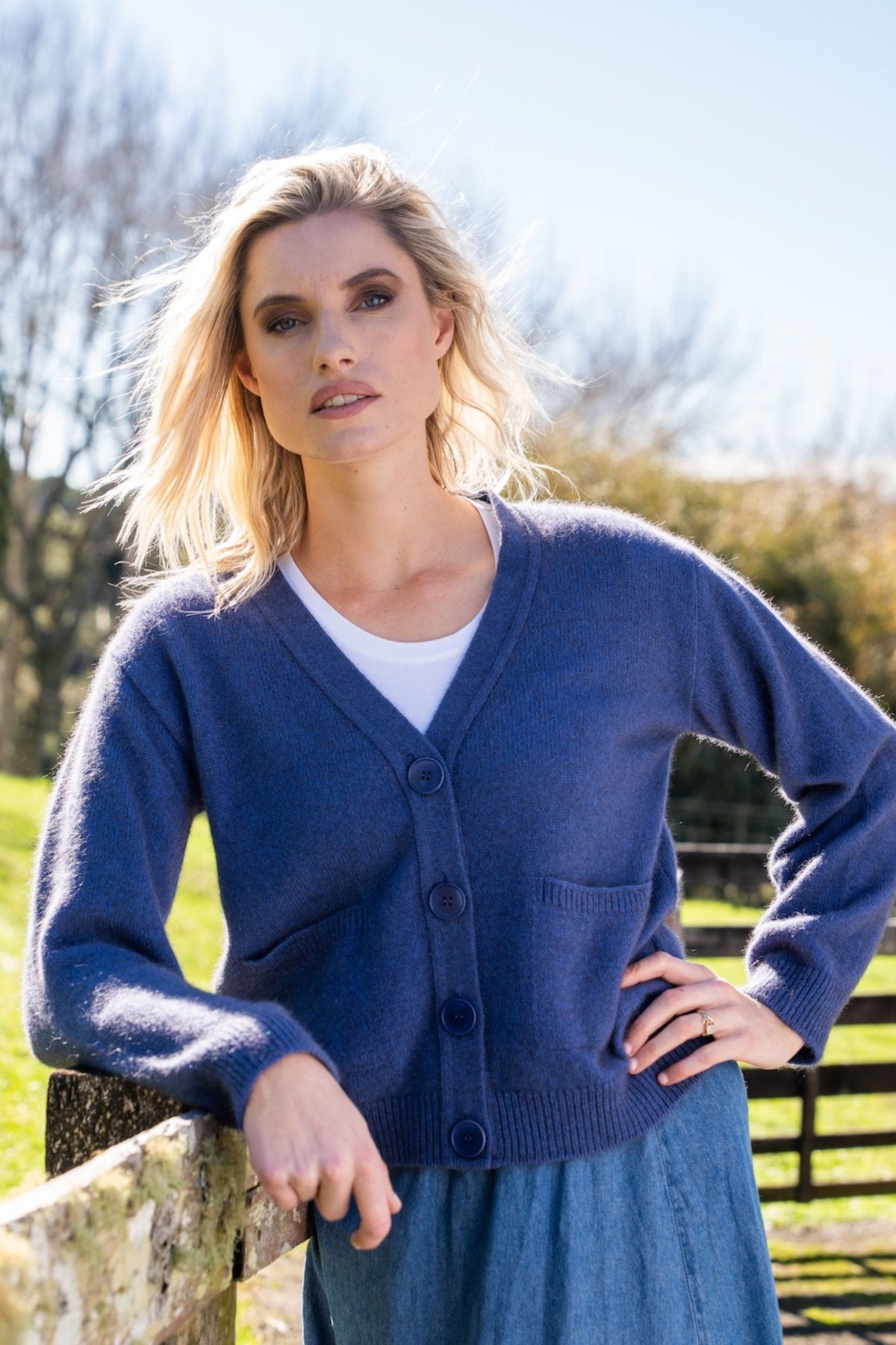 A woman wearing the Classic V Neck Possum Merino Cardigan in Periwinkle by Koru over a white top and blue skirt, standing outdoors near a wooden fence with trees and grass in the background.
