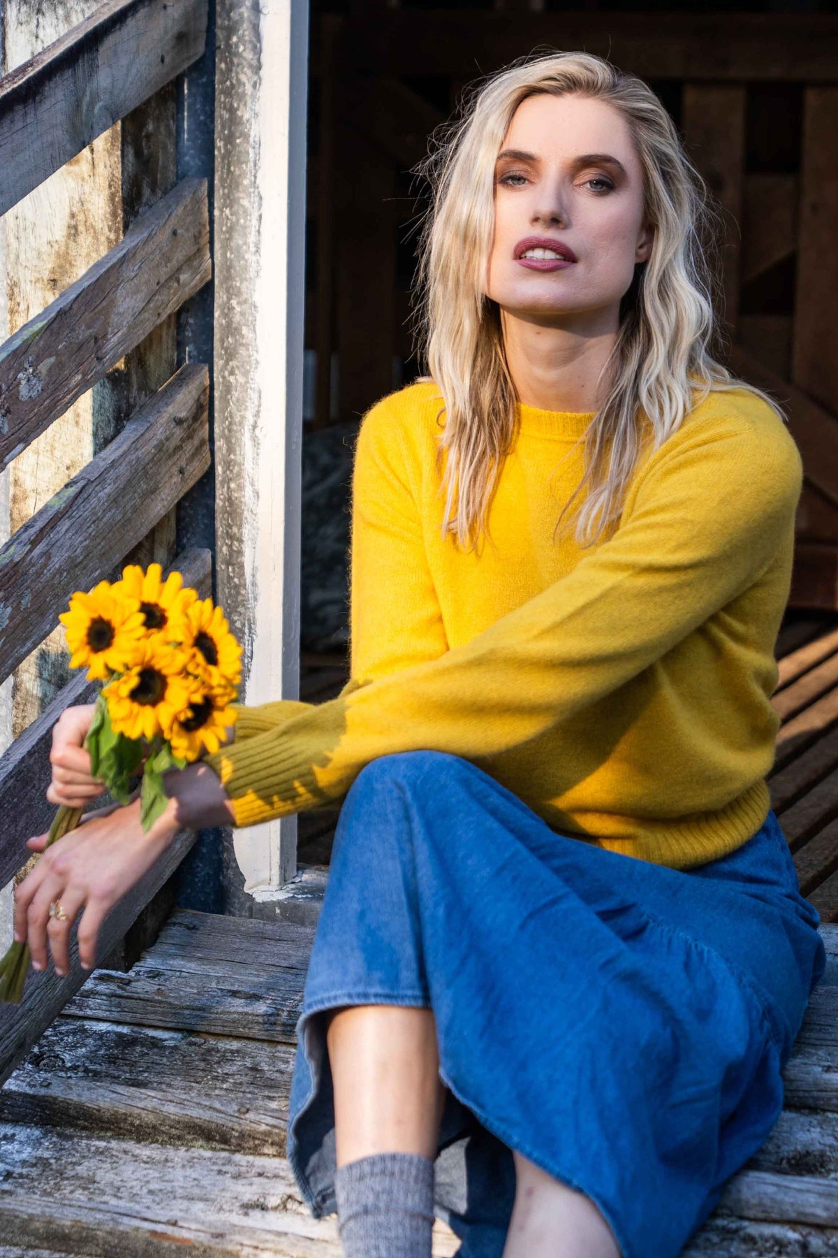A woman sits on rustic wooden steps holding a bouquet of sunflowers, wearing the Classic Crew Neck Possum Merino Jumper in Kowhai by Koru and a blue denim skirt.