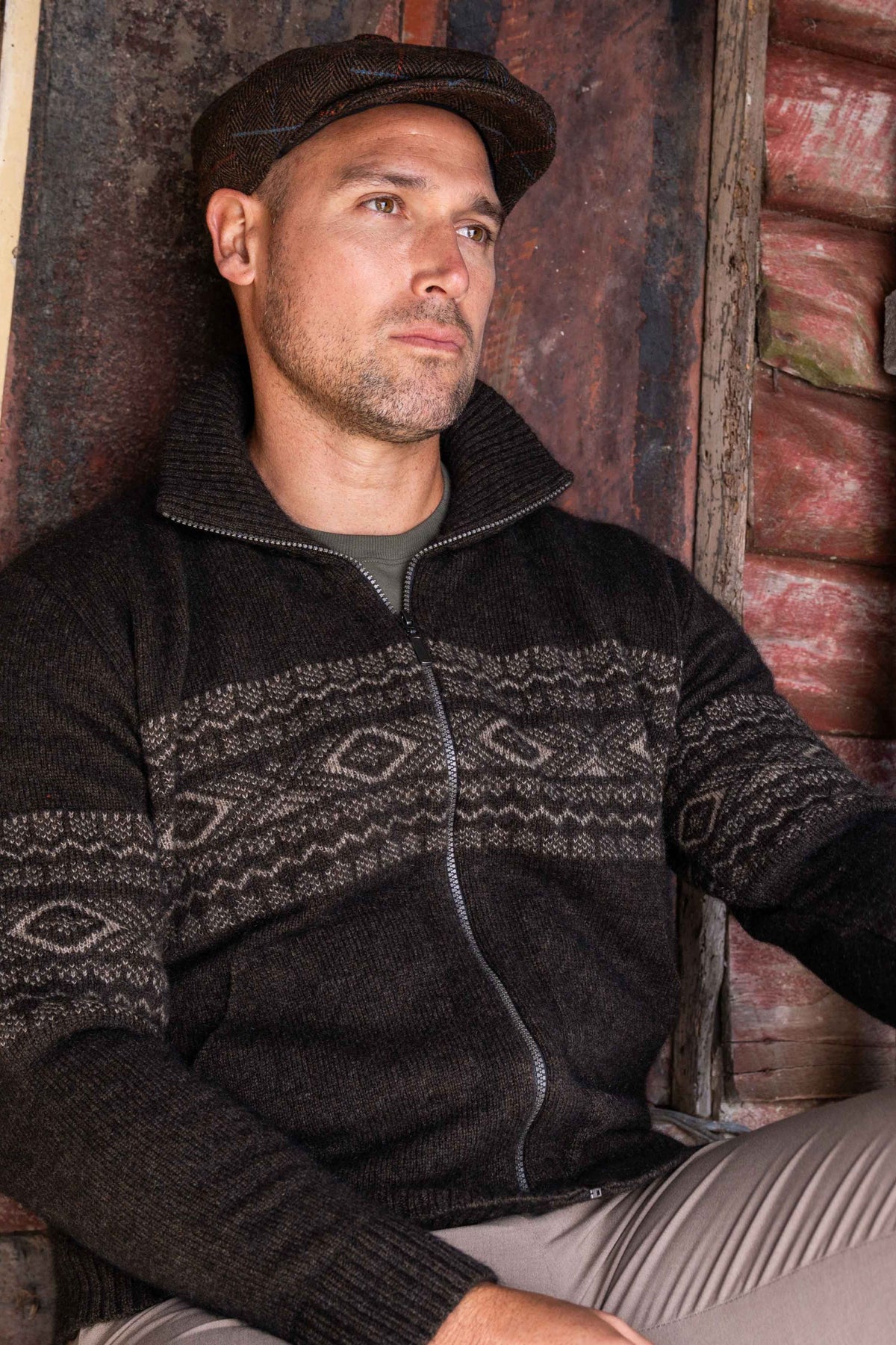 A man wearing the Fairisle Zip Possum Merino Jacket in Espresso/Mocha by Koru with a gray geometric pattern across the chest and sleeves, paired with a dark flat cap and light gray pants, leaning against a rustic wooden wall.