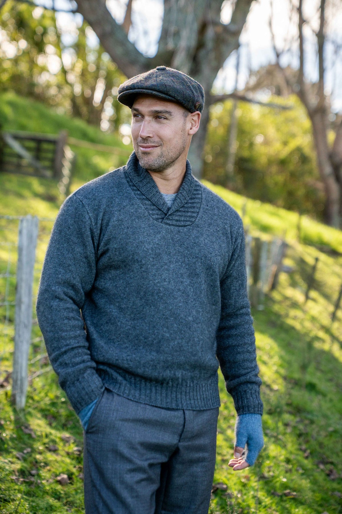 A man wearing the Shawl Collar Possum Merino Jumper in Grey by Koru, matching flat cap, and trousers, standing outdoors on a grassy hill with trees in the background.