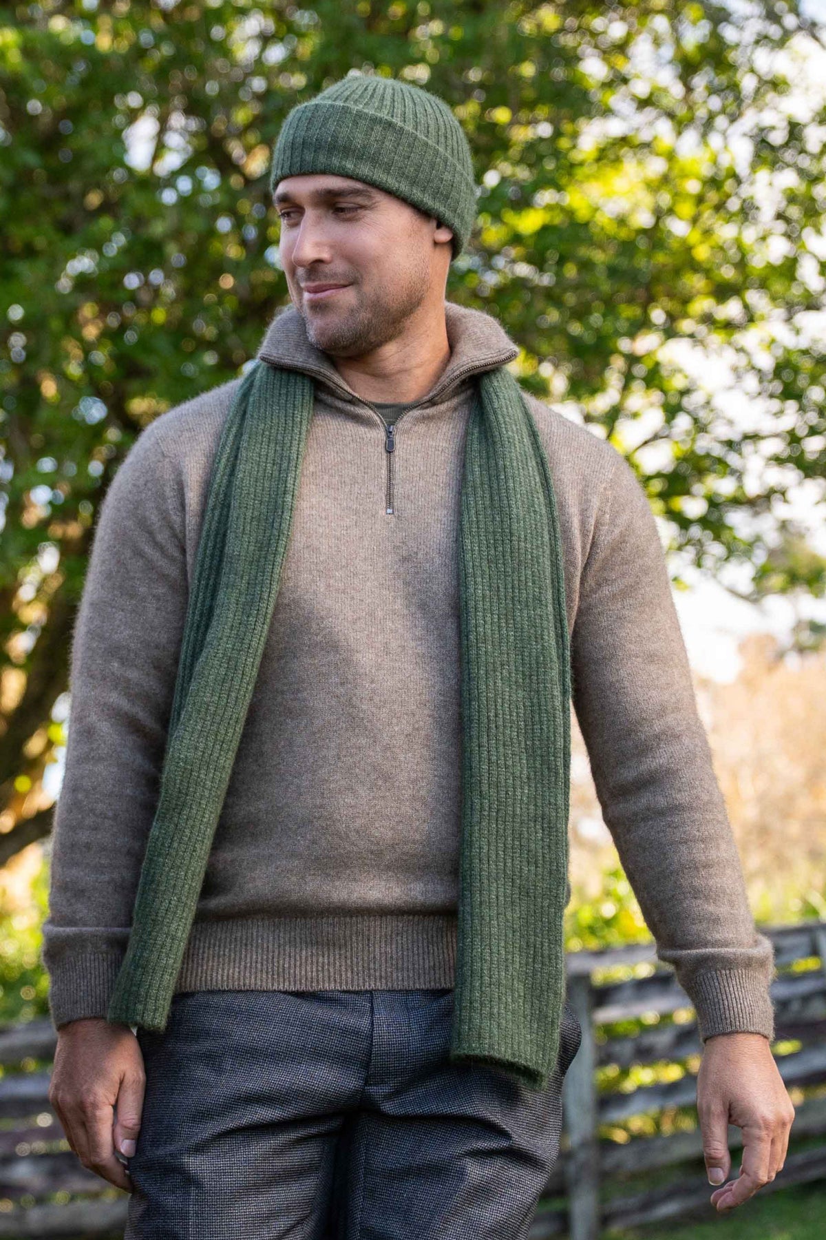 A man wearing a green knit beanie, a long green ribbed scarf, and the Everyday Quarter Zip Possum Merino Jumper in Mocha by Koru, standing outdoors in front of a blurred green tree background.