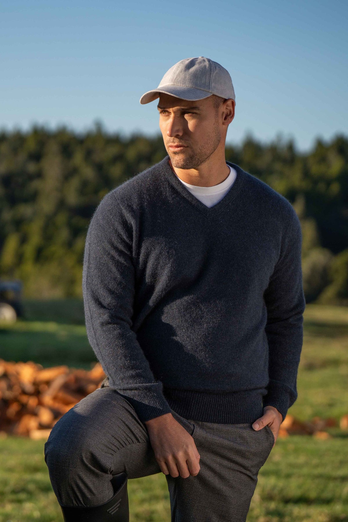 A man wearing the Classic V Neck Possum Merino Jumper in Indigo by Koru, gray pants, a light gray baseball cap, and a white undershirt, standing outdoors in a grassy field with trees and a blue sky in the background.