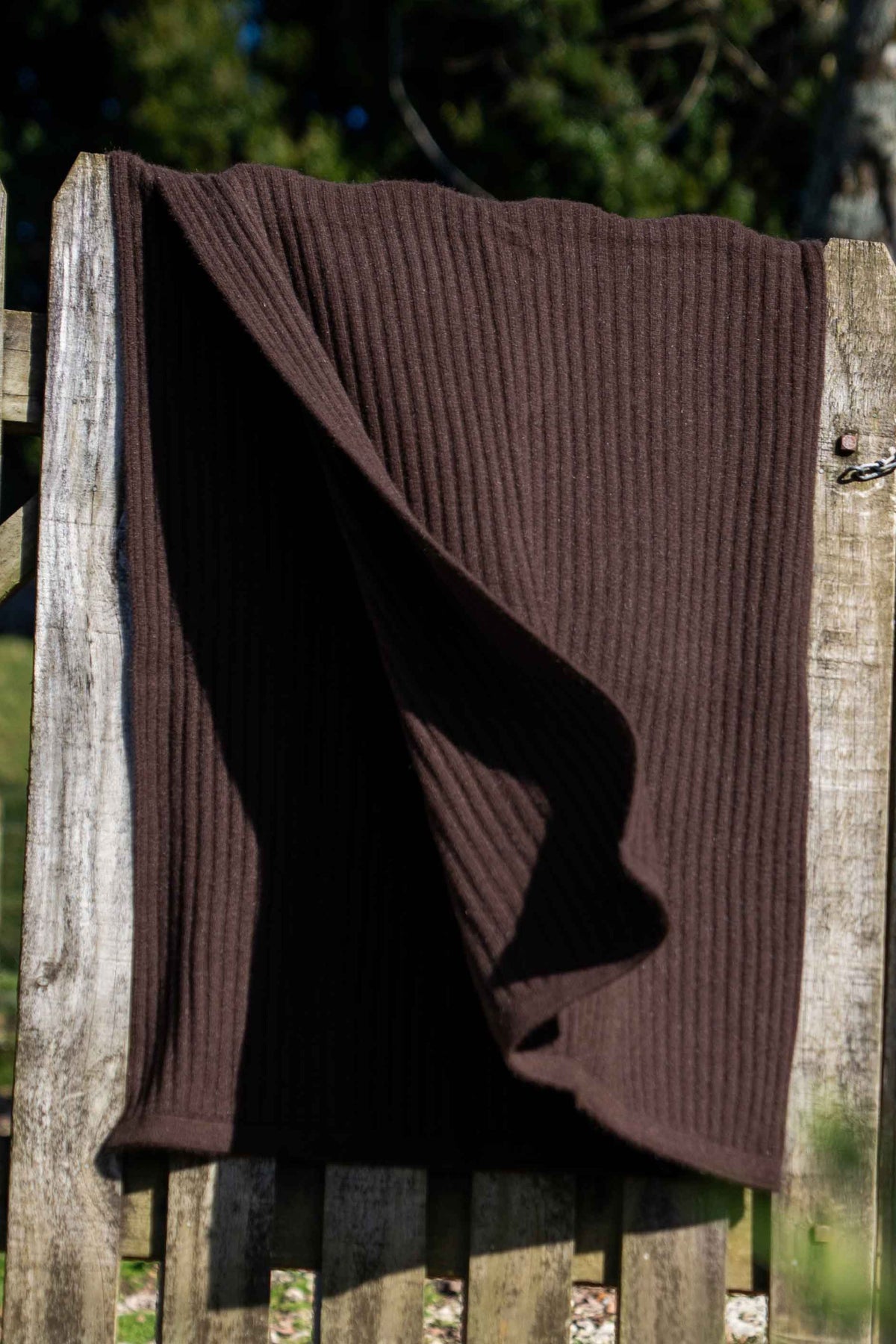 The Ribbed Possum Merino Throw in Chocolate by Koru draped over a weathered wooden picket fence in an outdoor garden setting.