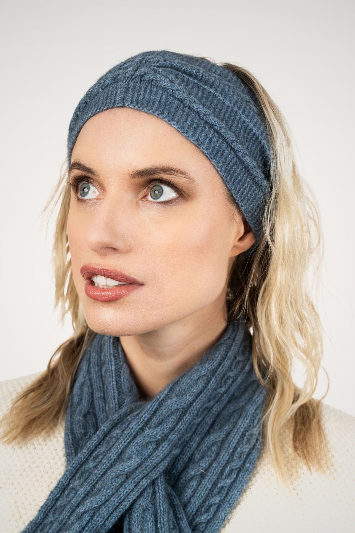 A woman wearing the the Cable Possum Merino Headband in Riviera by Koru and matching scarf, looking to the side against a light gray background.