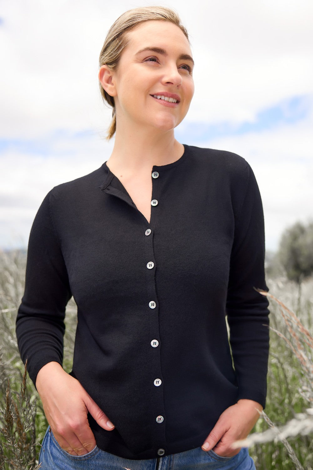 Woman wearing the Classic Merino Button Cardigan in Black by Royal Merino, standing outdoors with a blurred natural background
