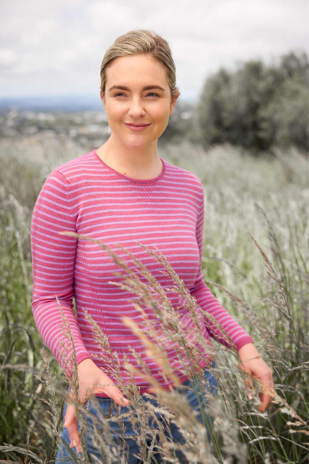 Woman wearing the Tuck Stitch Stripe Merino Jumper in Azalea/Lavender by Royal Merino, standing in a field with tall grass and trees in the background.