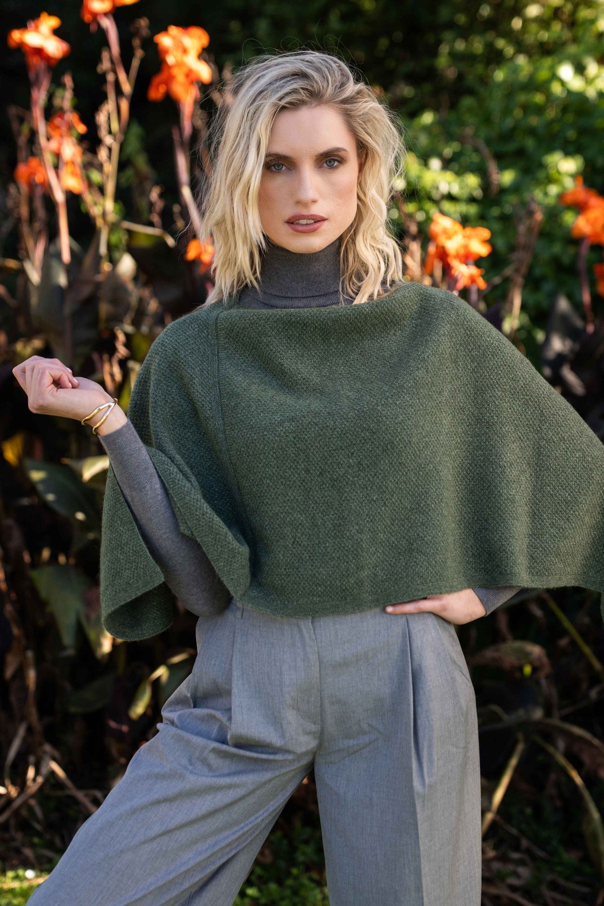 A woman wearing the Moss Stitch Possum Merino Poncho in Fern by Trilogy over a gray turtleneck and light gray wide-leg trousers, posing outdoors in front of a background of green foliage and orange flowers.