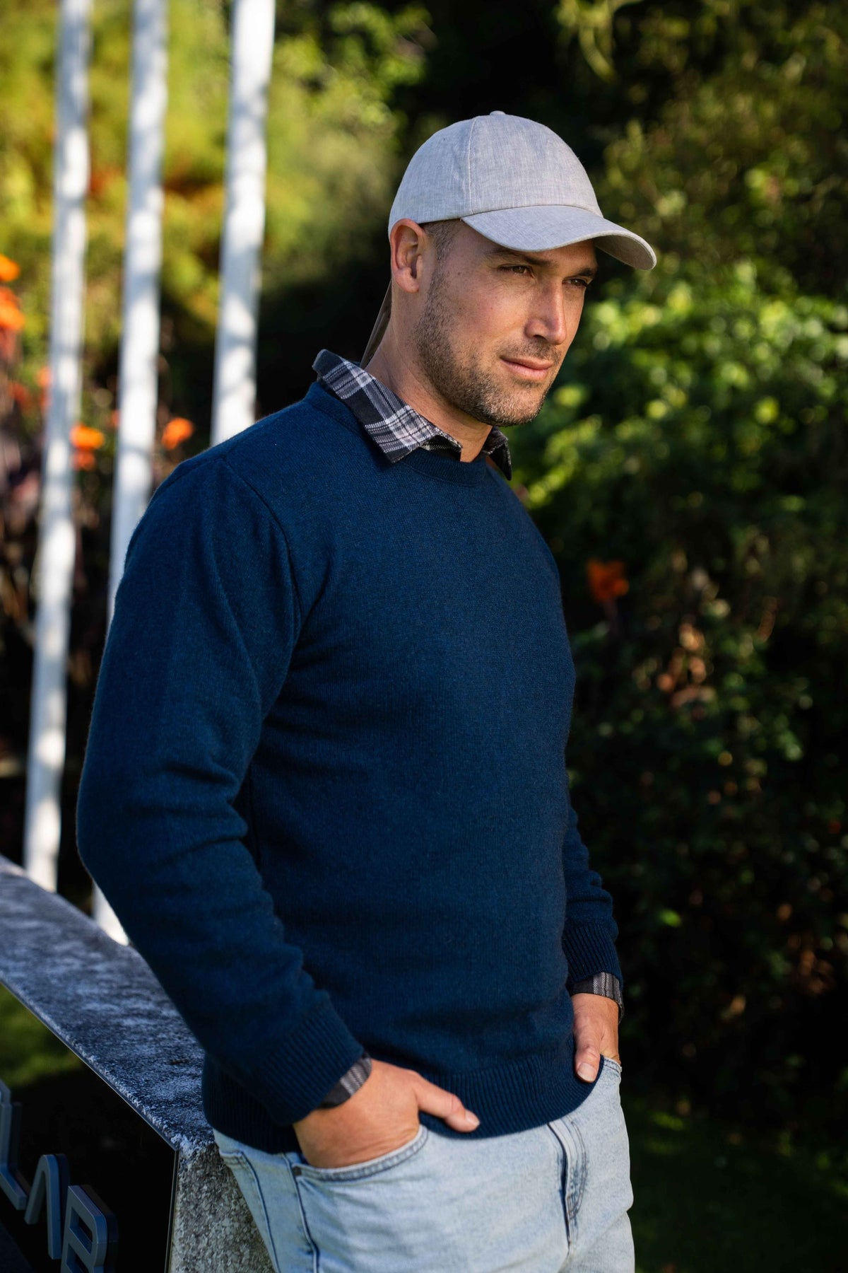 A man wearing the the Classic Possum Merino Crew Neck in Admiral by Trilogy over a plaid collared shirt, a light gray baseball cap, and light blue jeans, standing outdoors with trees in the background.