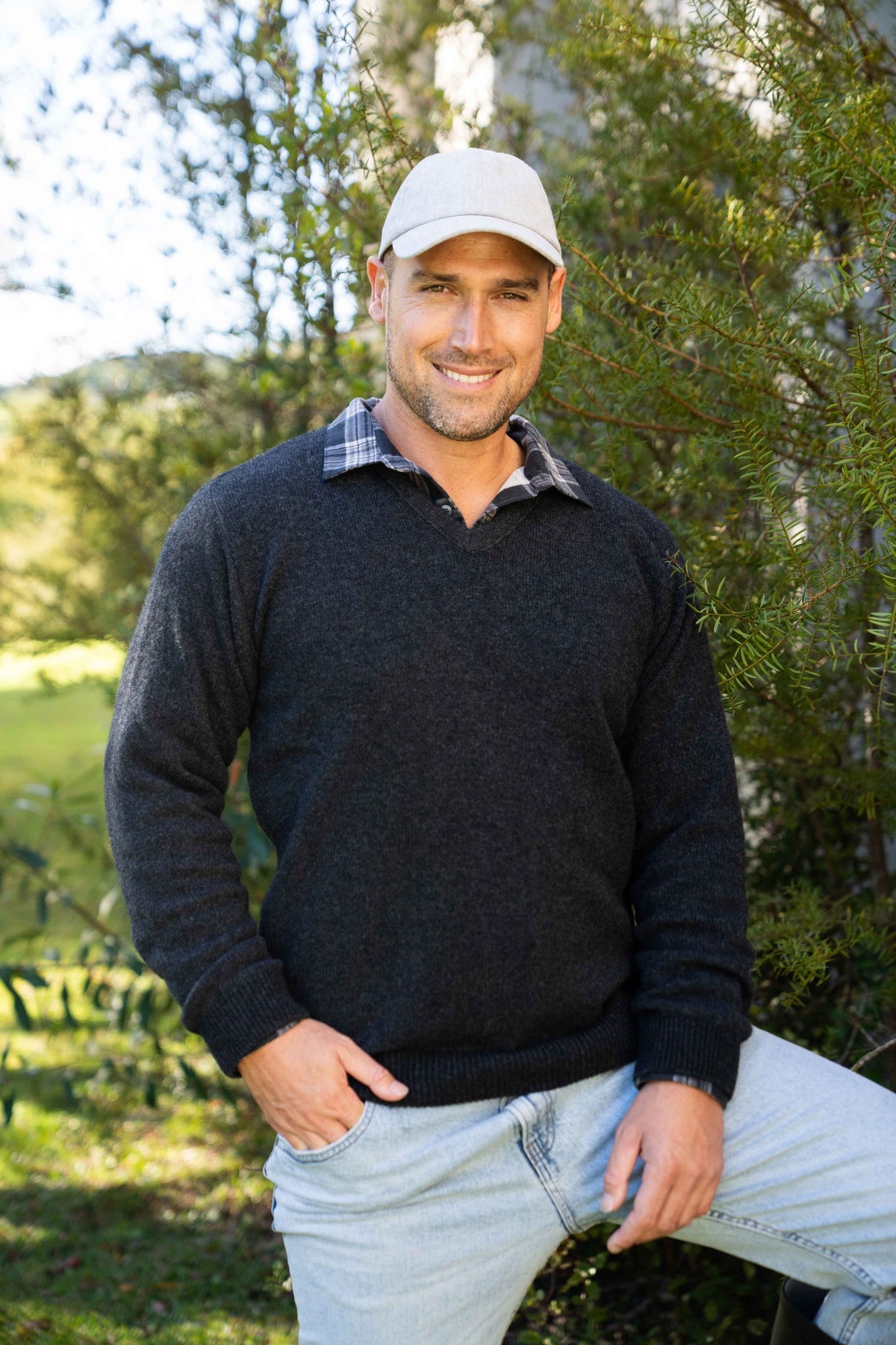 A man wearing the V Neck Possum Merino Jumper in Black Marl by Trilogy over a plaid collared shirt, a light gray baseball cap, and light blue jeans, standing outdoors in front of green trees.