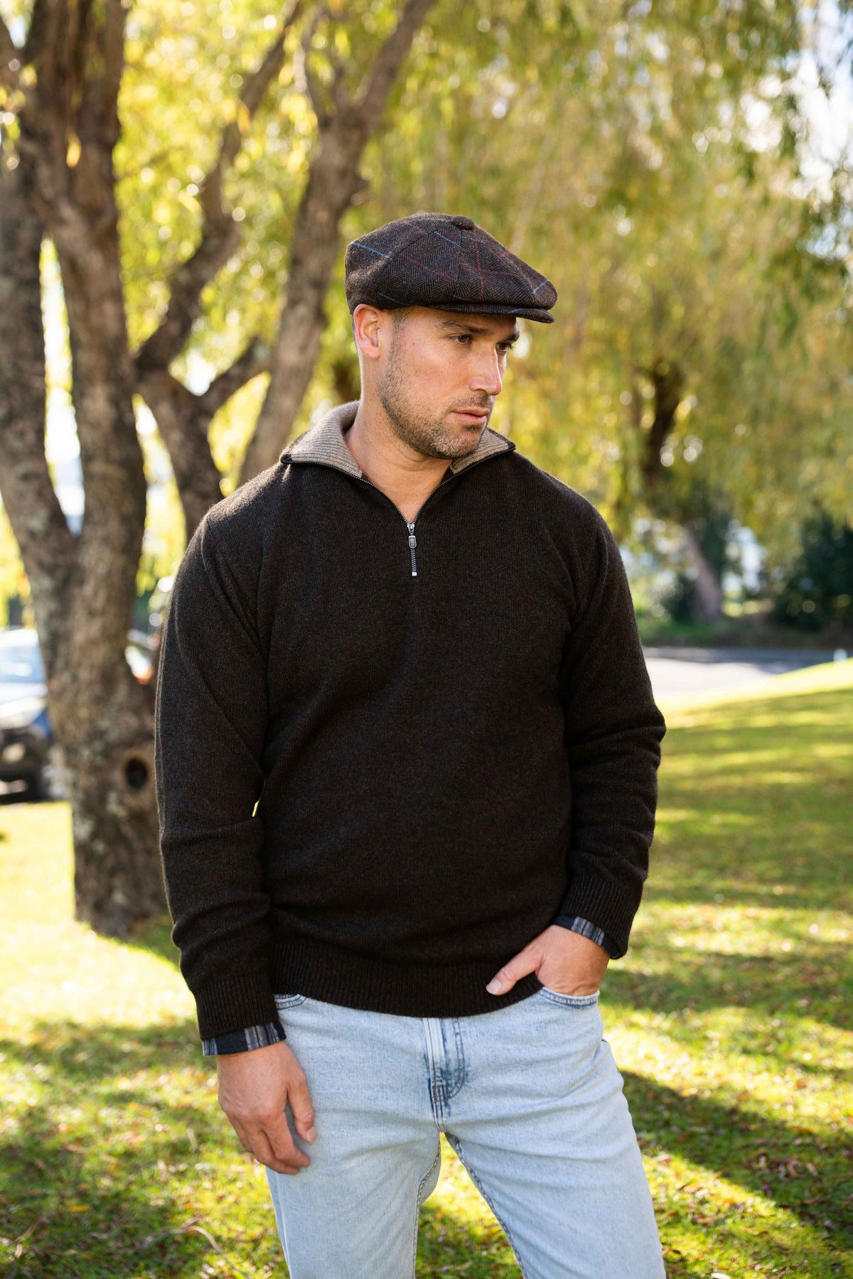 A man wearing the Zip Collar Possum Merino Jumper in Cocoa/Mocha by Trilogy, a dark flat cap, and light blue jeans, standing outdoors in a park with trees and grass in the background.