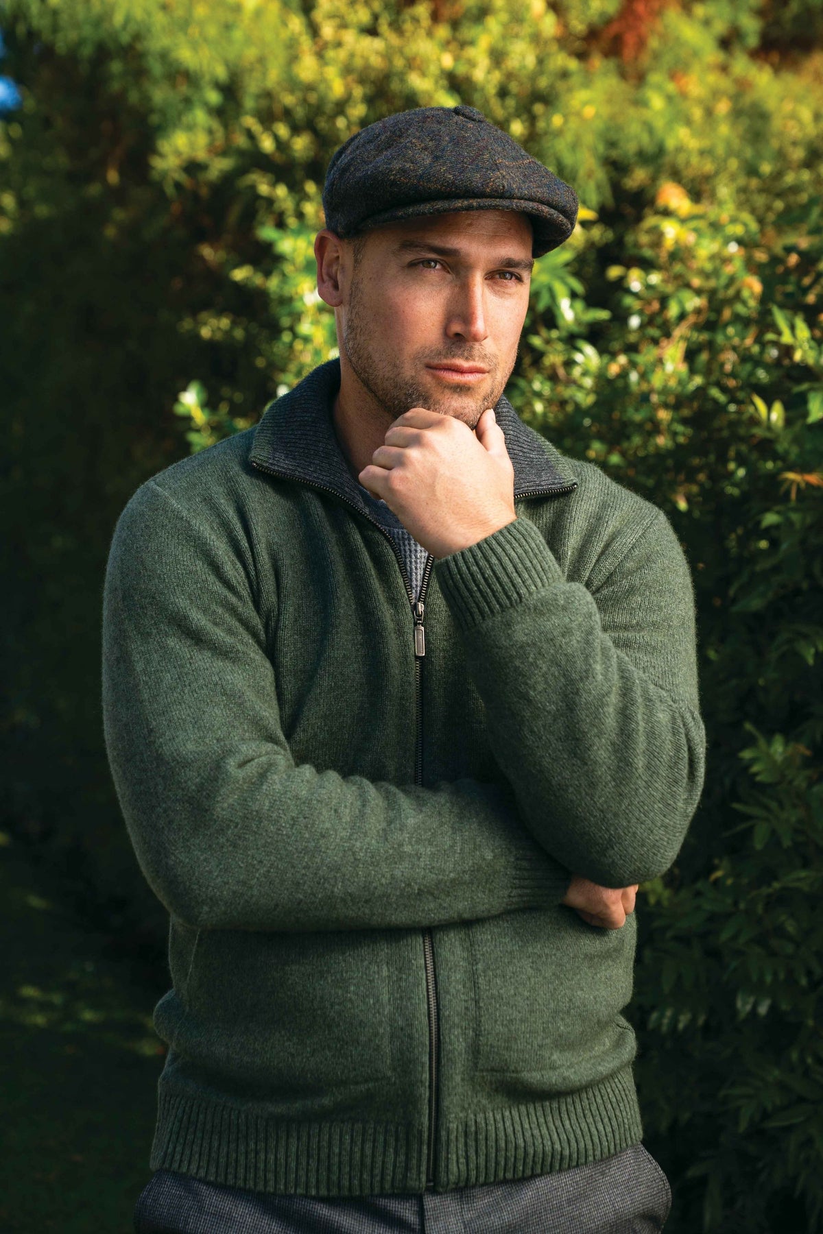 A man wearing the Zip Possum Merino Jacket in Fern/Grey by Trilogy and a dark flat cap, standing outdoors in front of a green hedge