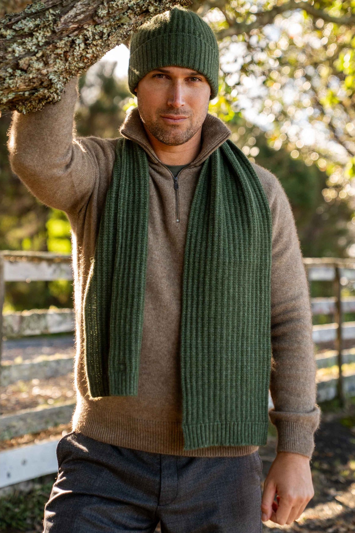 A man wearing the Ribbed Possum Merino Scarf and Beanie in Moss by Koru over a brown zip-up sweater, standing outdoors near a tree trunk and fence.