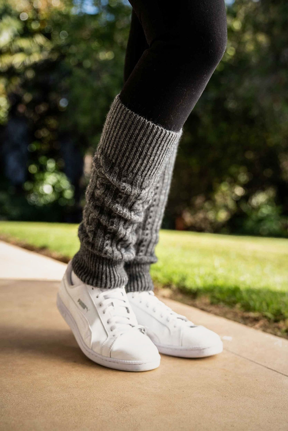 Person wearing Cable Possum Merino Leg Warmers in Grey by Koru with white sneakers on a blurred outdoor background