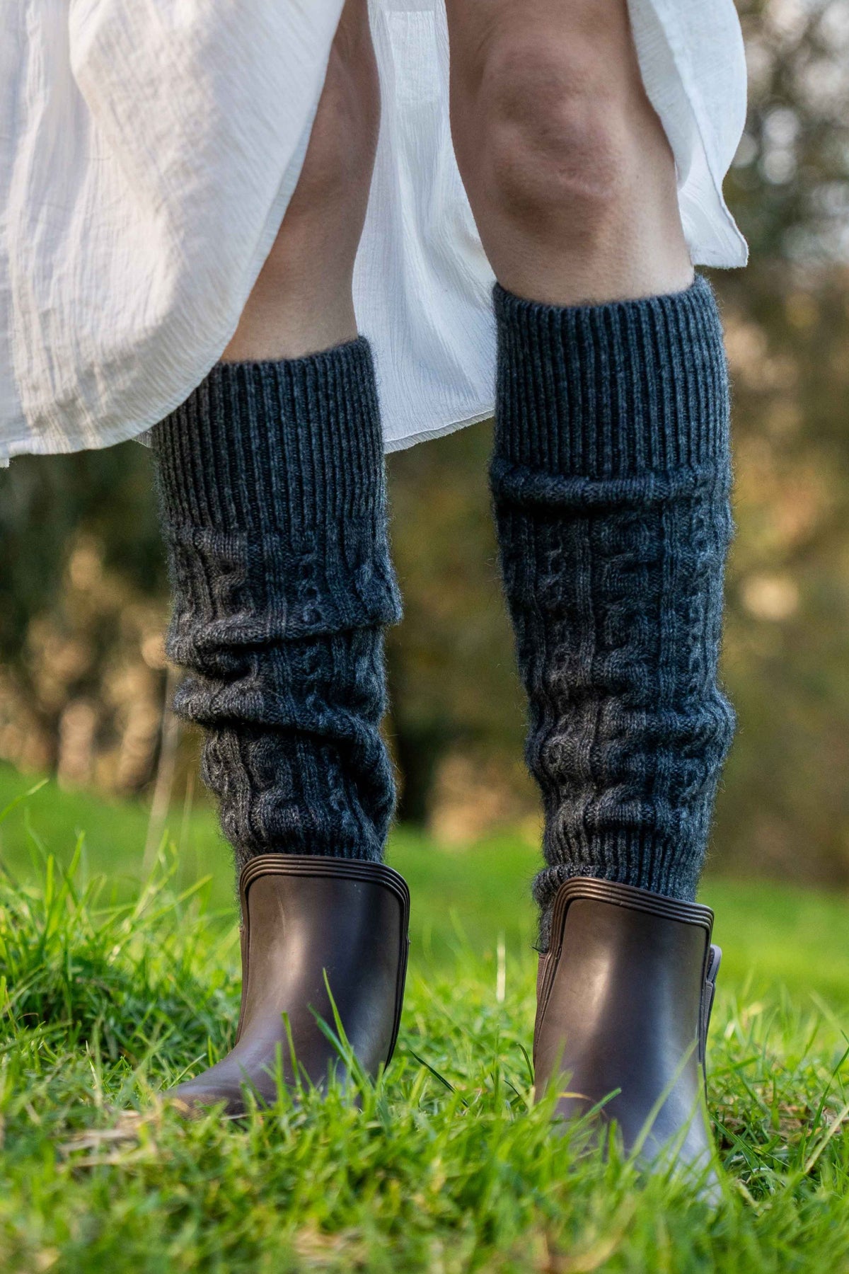 A close-up of a woman's legs wearing the Cable Possum Merino Leg Warmers in Grey by Koru and dark brown boots on a green grassy lawn.