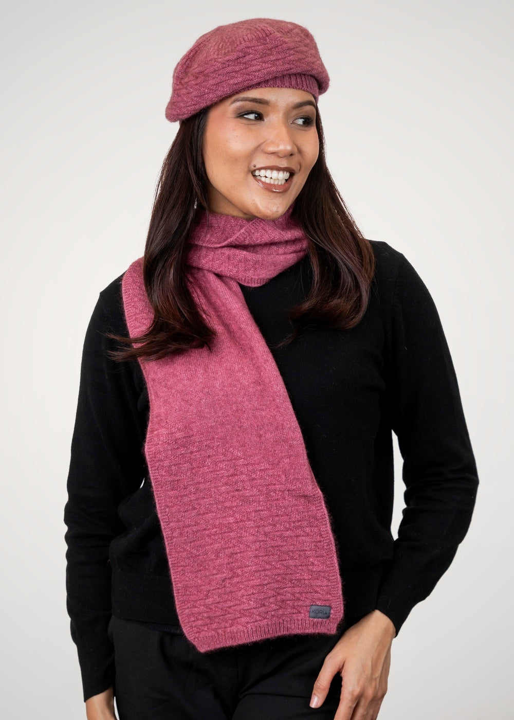 Woman wearing the Zig Zag Possum Merino Scarf in Rose by Koru with matching hat on a white background