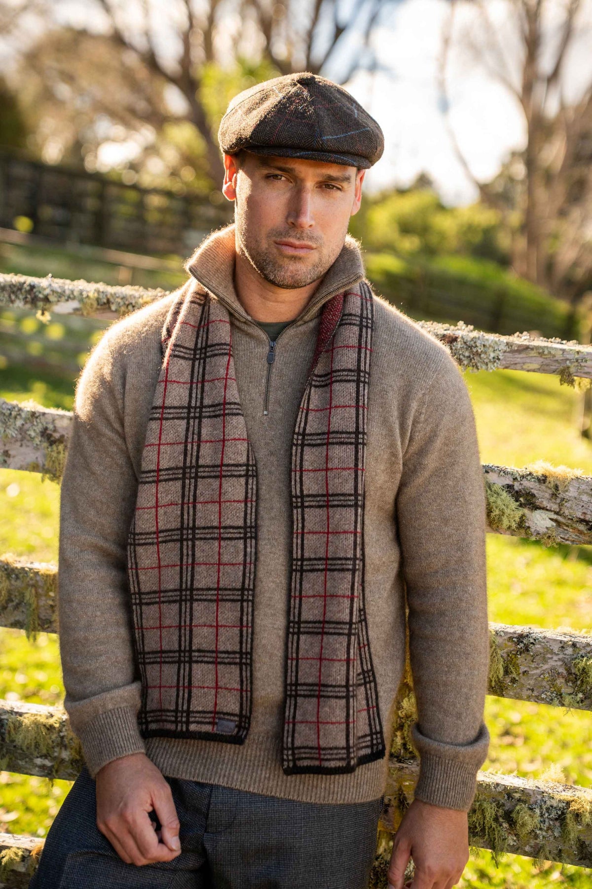 A man wearing a brown flat cap, a taupe knit zip-up sweater, and the Tartan Possum Merino Scarf in Mocha/Espresso/Rata by Koru, standing outdoors in front of a rustic wooden fence.