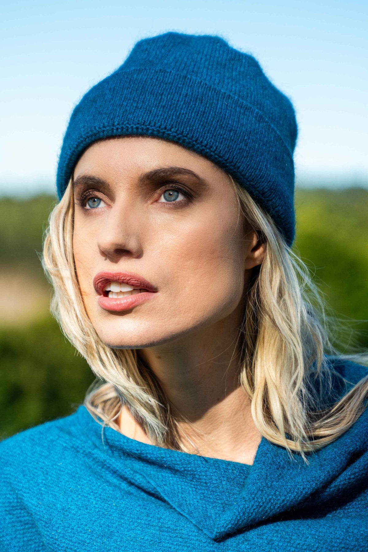 A close-up portrait of a woman wearing the Plain Possum Merino Beanie in Pacific by Koru and a matching poncho, photographed outdoors with a blurred green and blue background.