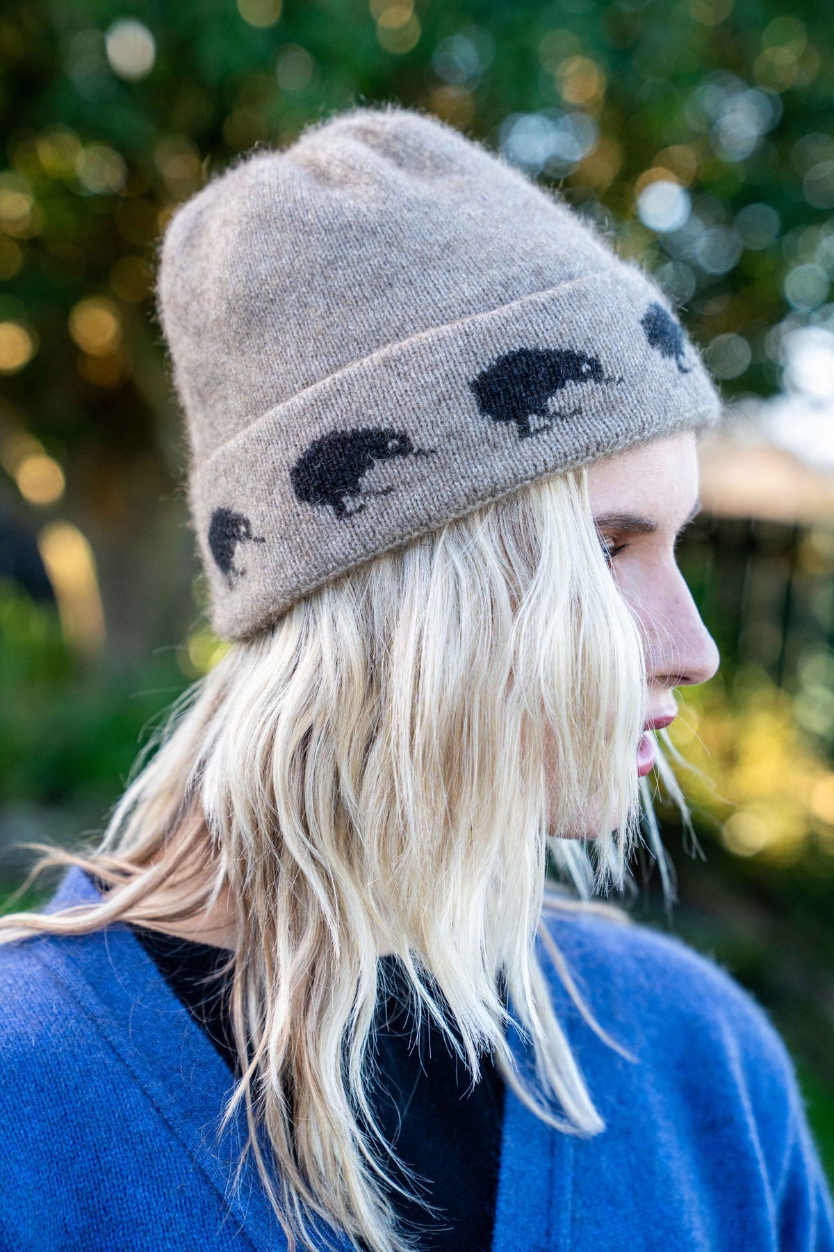 A woman in profile wearing the Kiwi Possum Merino Beanie in Mocha by Koru, photographed outdoors with a blurred green background.