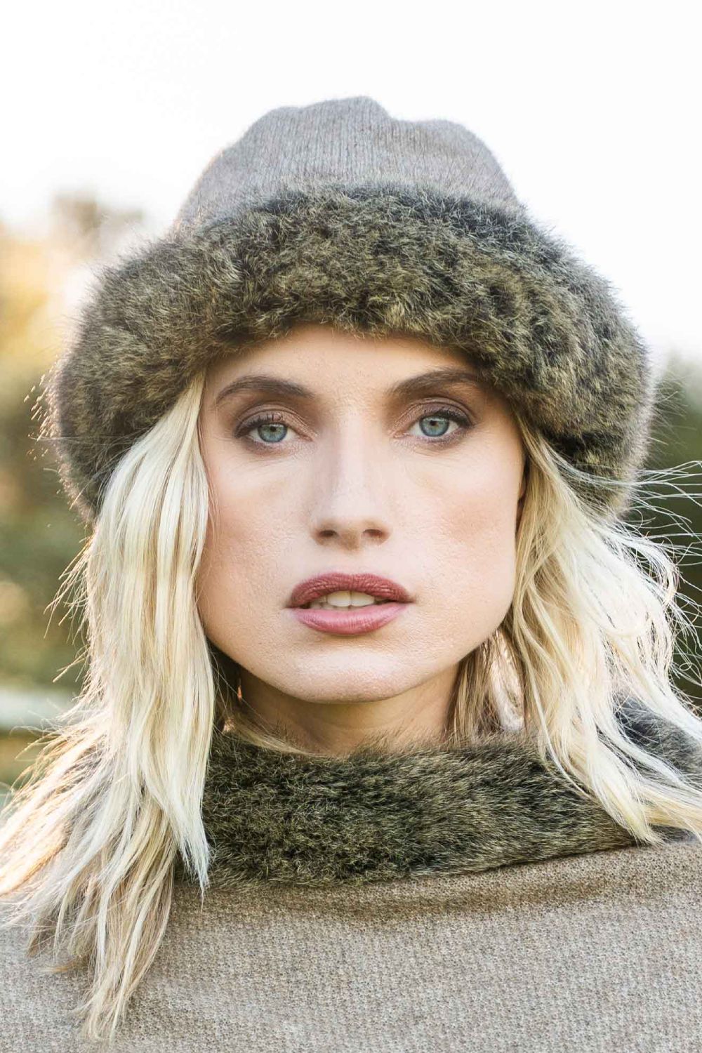 A close-up portrait of a woman wearing the Fur Trim Possum Merino Beanie in Mocha/Snowtip by Koru, with matching Poncho set against a soft, blurred outdoor background.