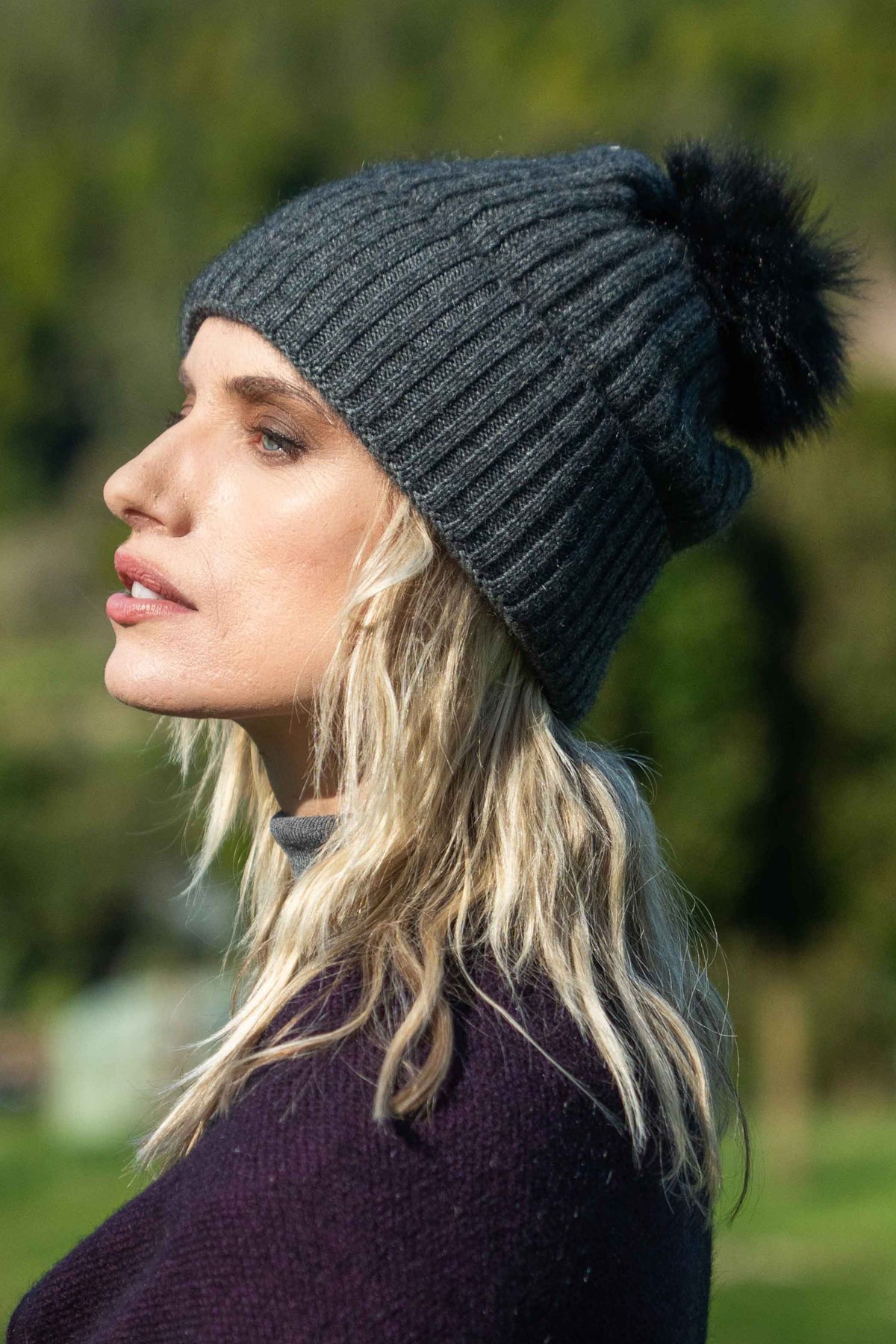 A woman in profile wearing the Fur Pom Pom Possum Merino Beanie in Grey by Koru, set against a blurred green outdoor background.