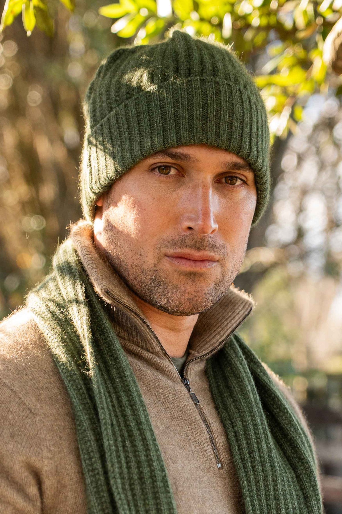A man wearing the Ribbed Possum Merino Beanie in Moss by Koru and matching scarf, photographed outdoors with blurred trees and sunlight in the background.