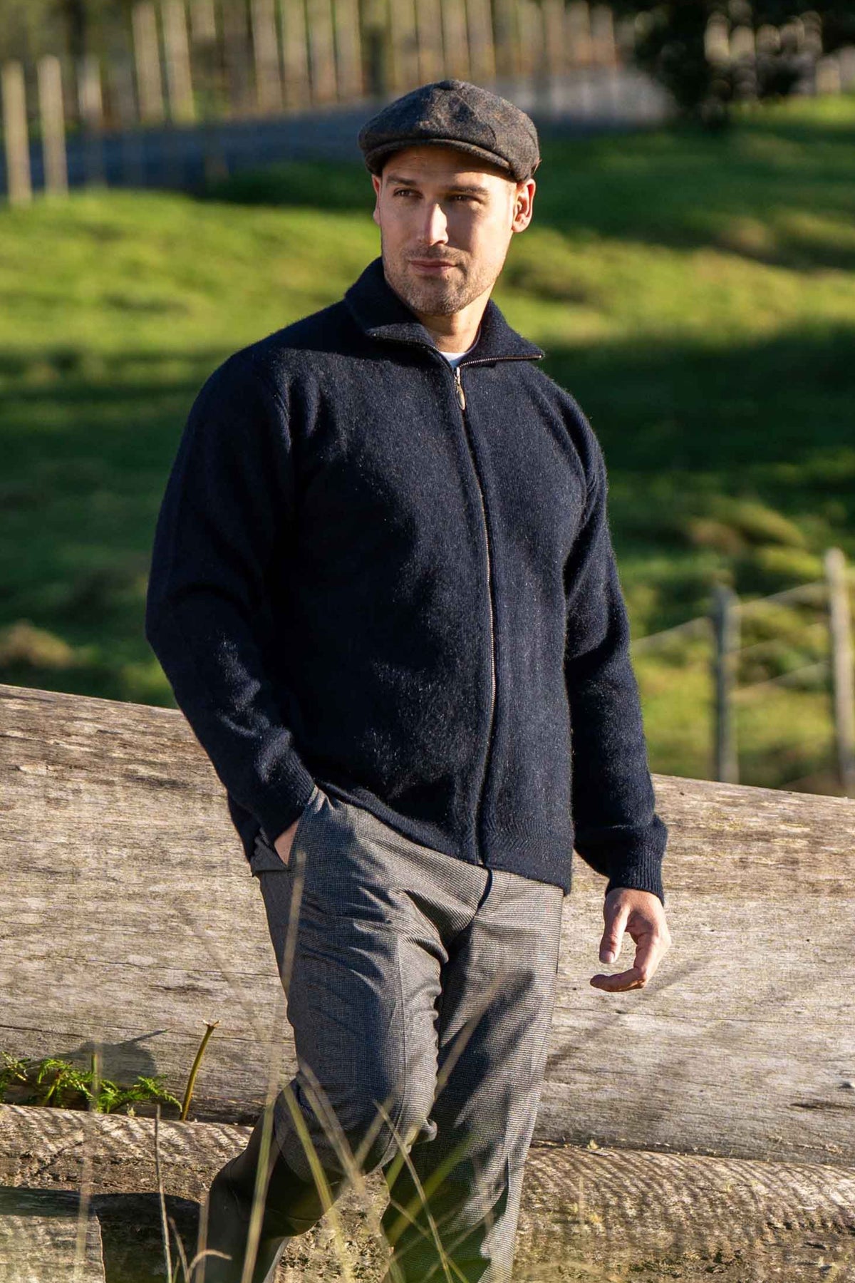 A man wearing the Zip Through Possum Merino Jacket in Navy by Koru, gray trousers, and a dark flat cap stands outdoors leaning against a large wooden log with a grassy field in the background.