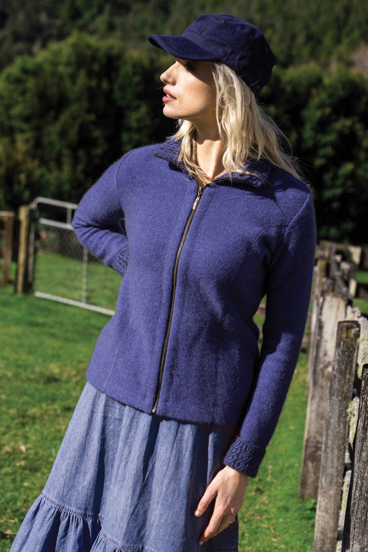 A woman wearing the Two Tone Possum Merino Jacket in Periwinkle/Black by Koru,  a matching blue cap, and a denim skirt, standing outdoors in a grassy field with a wooden fence and trees in the background.