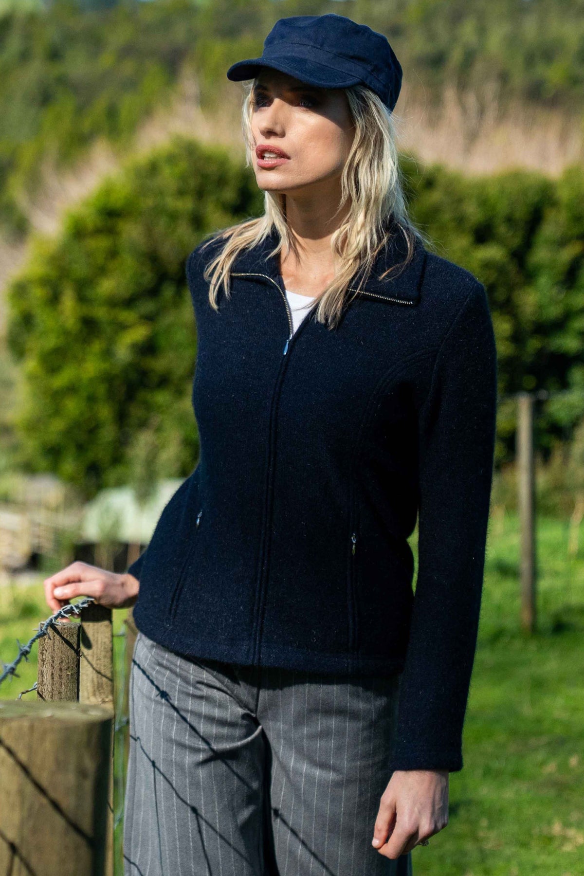A woman wearing the Shaped Zip Possum Merino Jacket in Navy by Koru, matching navy cap, and gray pinstripe trousers, standing outdoors in a grassy field.