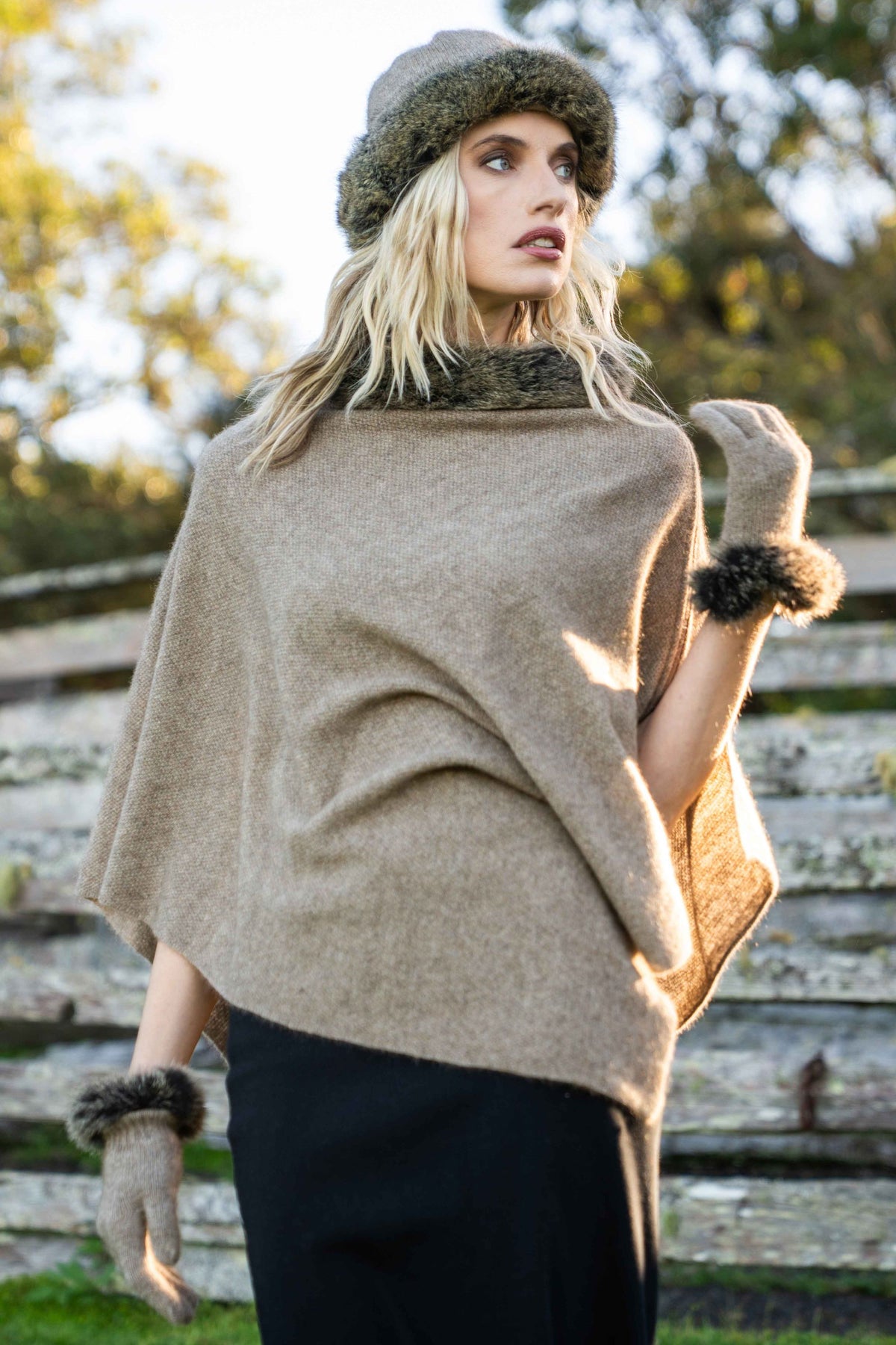 A woman wearing the the Fur Trim Possum Merino Poncho in Mocha/Snowtip by Koru, with matching gloves and beanie, standing outdoors in front of a rustic wooden fence.