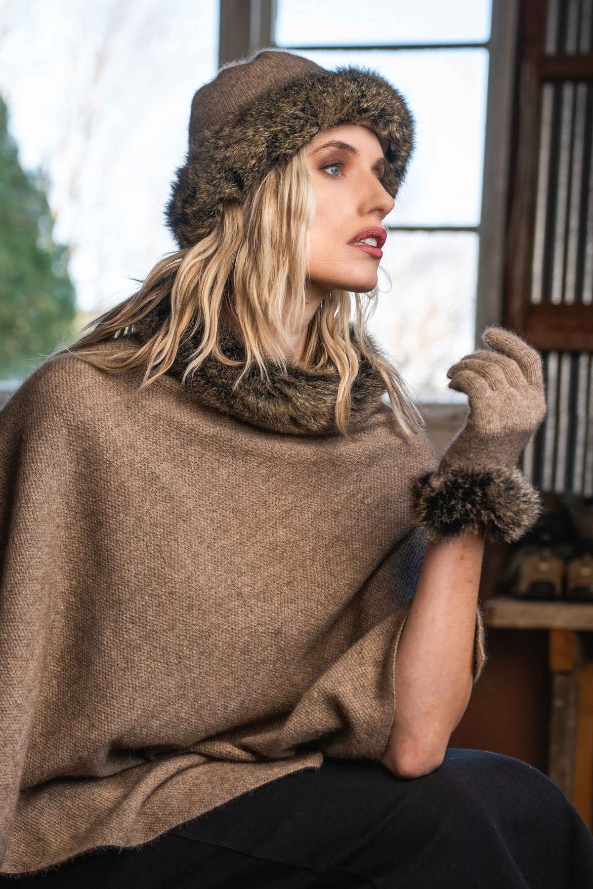 A woman wearing the the Fur Trim Possum Merino Collection  in Mocha/Snowtip by Koru, consisting of a poncho, gloves and beanie, with black pants, posed in front of a window.