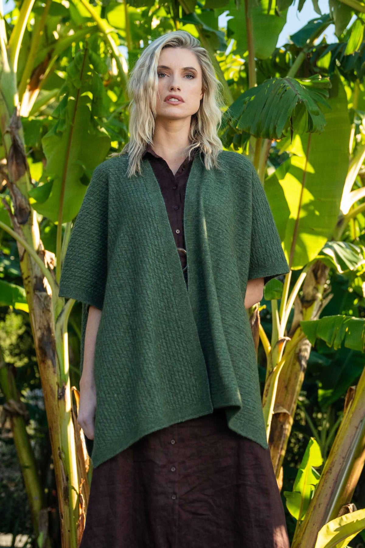 A woman wearing the Zig Zag Textured Possum Merino Cape in Moss by Koru  over a dark brown collared dress, standing outdoors in front of lush green banana leaves.